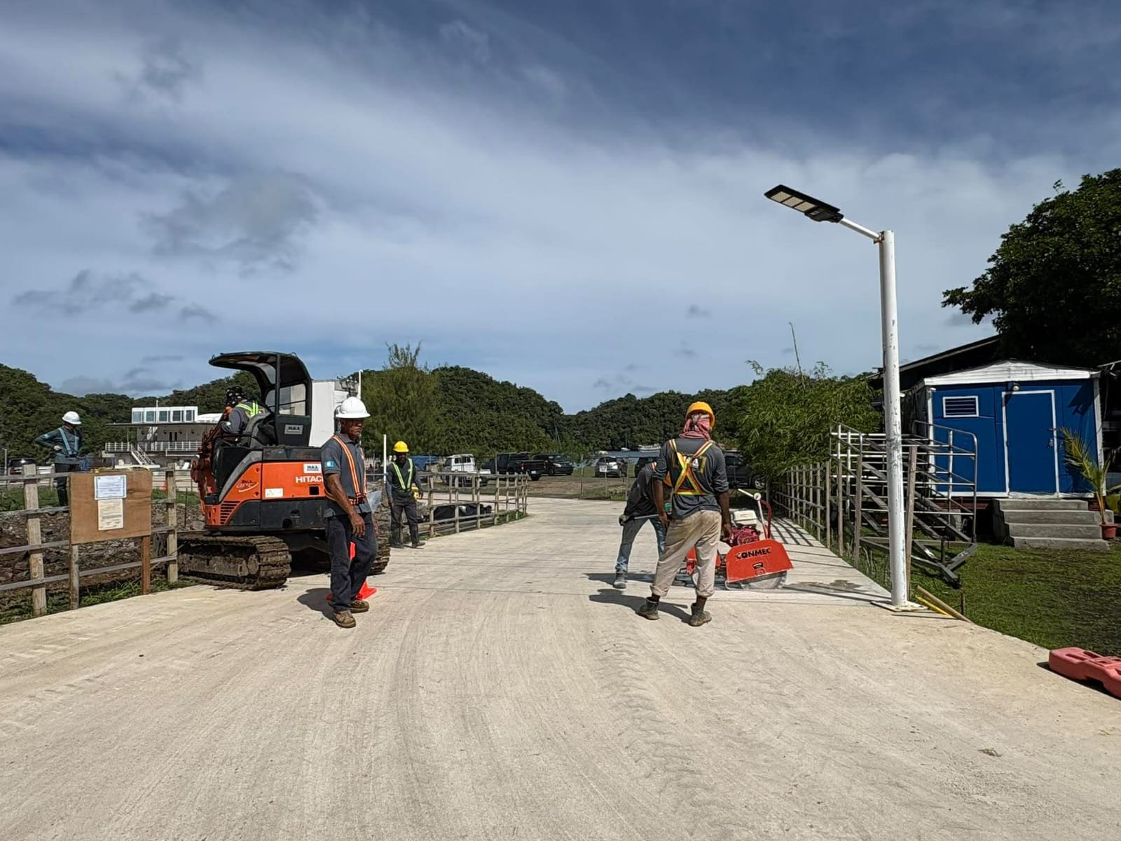 Construction workers installing a streetlight on a sidewalk with construction equipment and parked cars in the background.