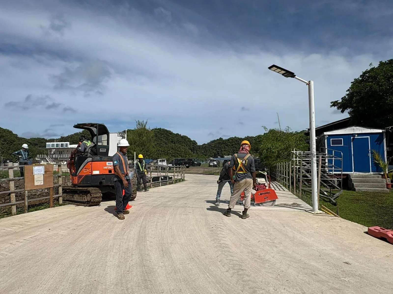 Construction workers on a dirt road with heavy machinery, a blue building, and trees in the background under a partly cloudy sky.