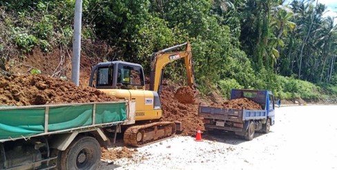 A construction site with a small yellow excavator digging into the dirt next to a blue truck, surrounded by green trees and bushes.