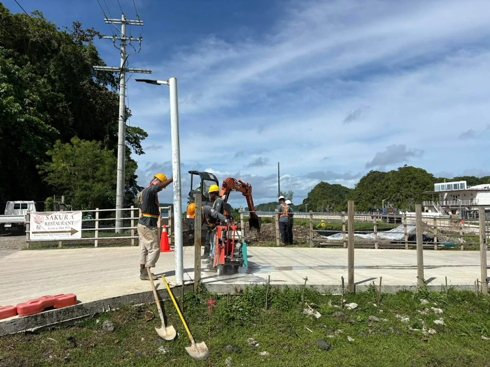 Construction workers wearing yellow helmets working near electrical poles and equipment outdoors on a partly cloudy day. There is a sign for Sakura restaurant in the background and a grassy area in the foreground.