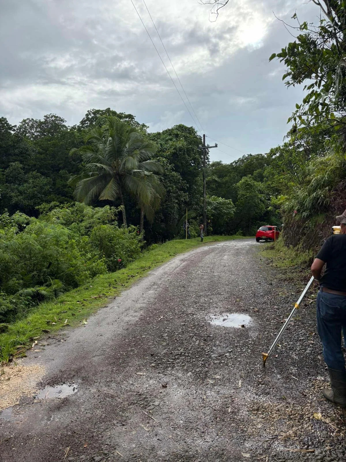 A person with a black shirt and blue pants is standing on a wet gravel road, holding a tool. There are two potholes filled with water on the road. The scene is surrounded by lush green trees and bushes, with a cloudy sky overhead and a red car parked on the side of the road.