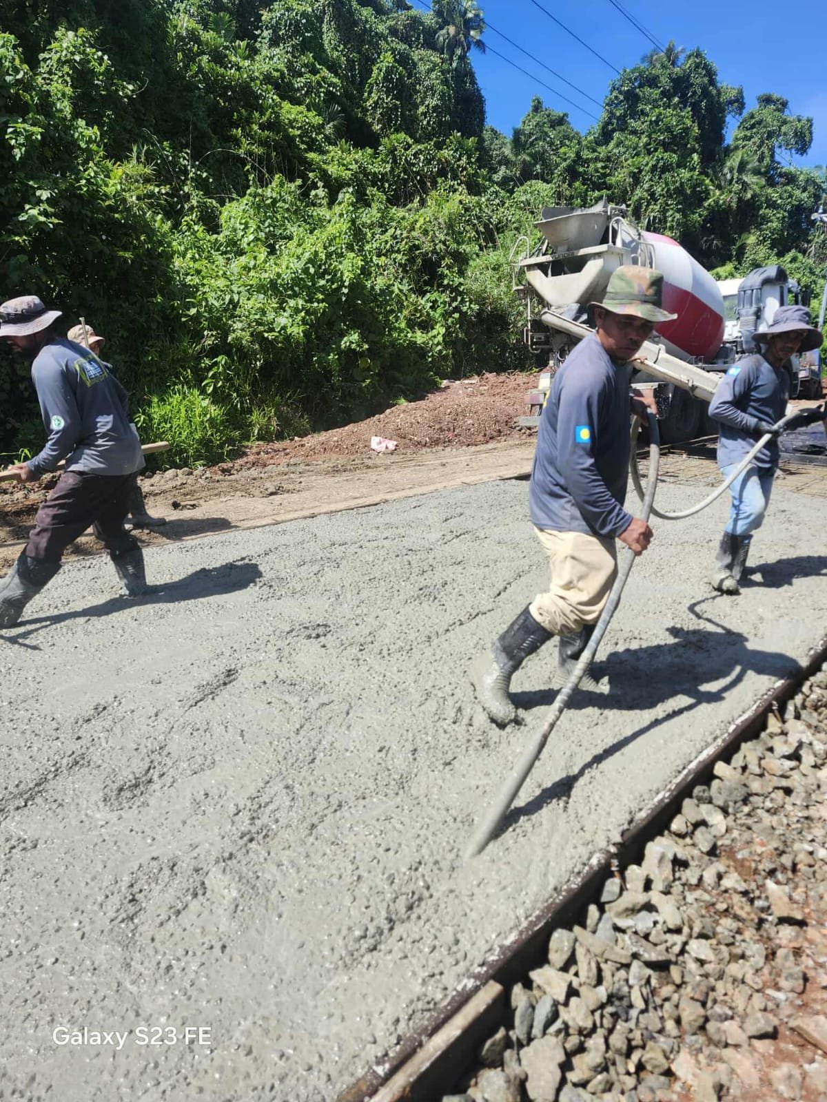 Construction workers laying concrete on a road with greenery in the background and power lines overhead.