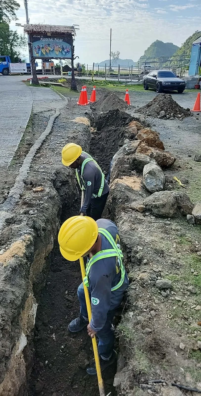 Two construction workers wearing yellow helmets and safety vests working in a trench on a construction site. The site has a road, orange safety cones, and a parked black car in the background with mountains in the distance.