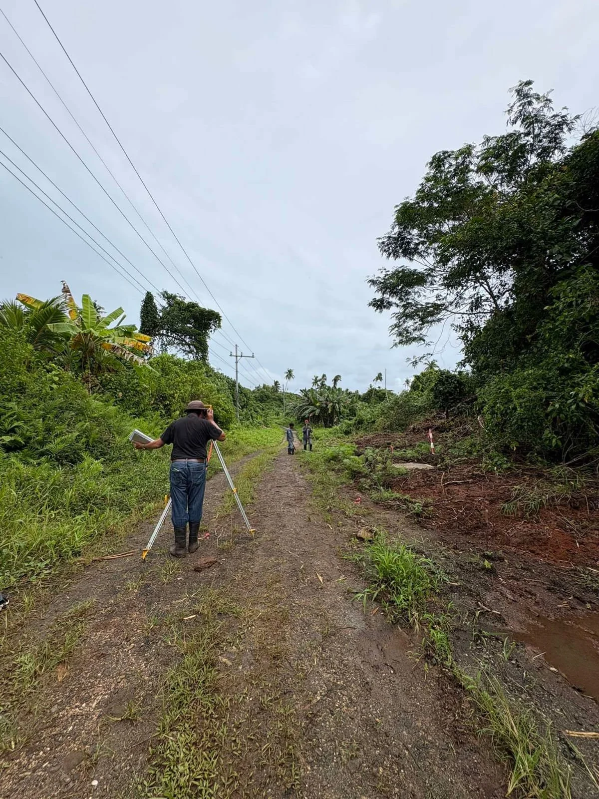 A man using surveying equipment on a dirt road surrounded by greenery with two people walking in the distance.