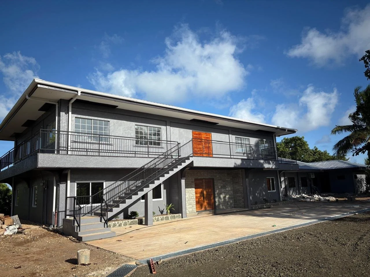 New gray two-story house under construction with stairs and unfinished driveway, blue sky, and trees in background.