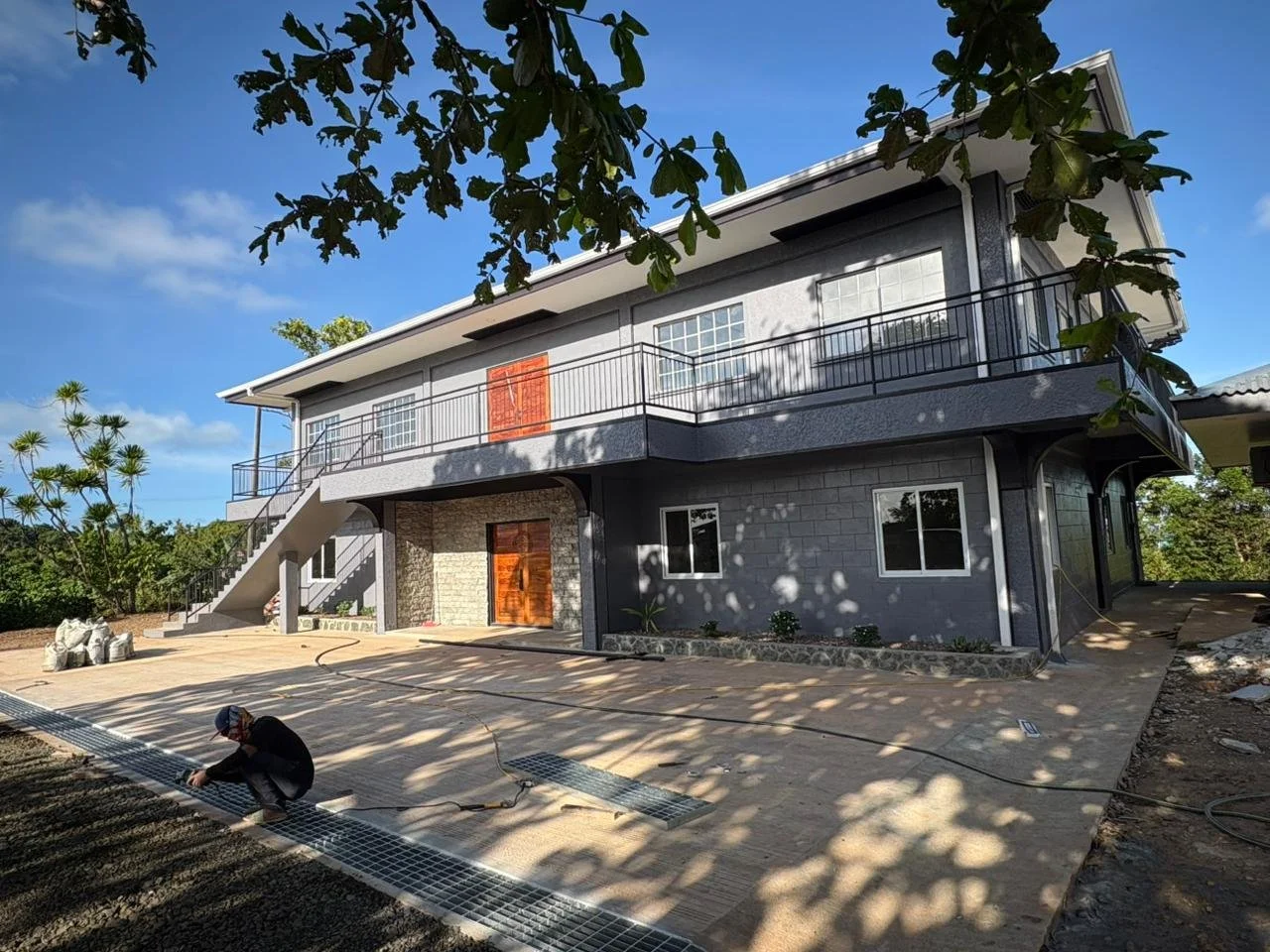 A two-story modern house with a gray exterior, white trim, and wooden accents, surrounded by trees under a blue sky. A person is working on the paved area in front of the house.