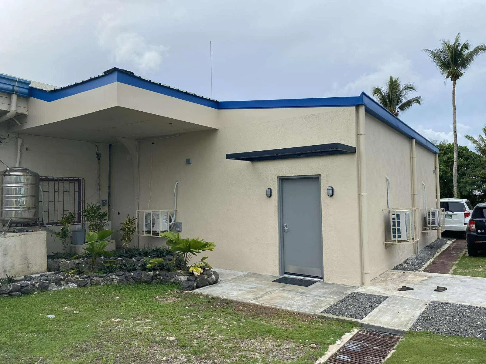 Exterior of a beige building with a blue roof, featuring a grey door, two wall-mounted air conditioners, and outdoor lighting. There are small plants and landscaping near the entrance, and the parking area has cars and palm trees in the background.