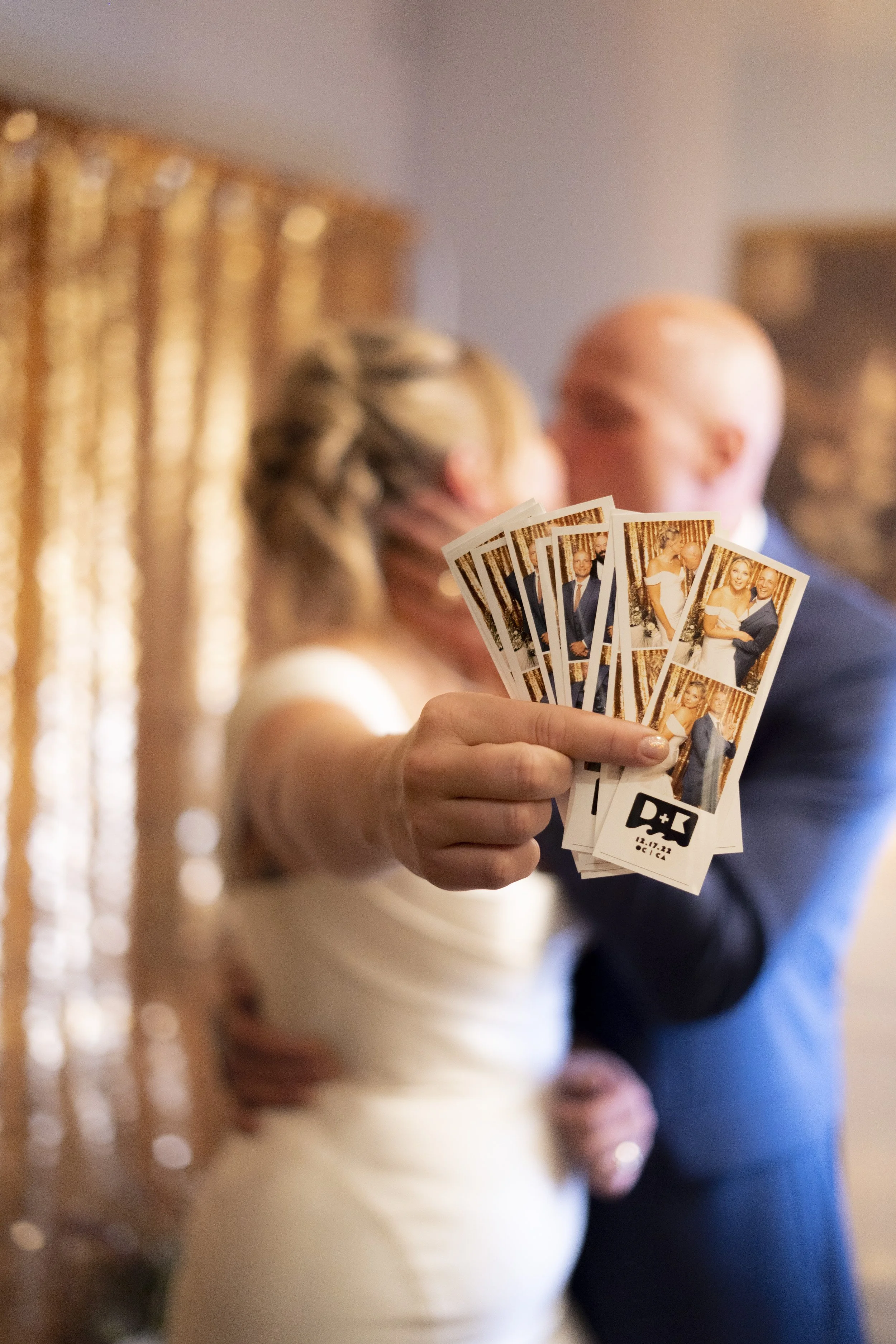 A woman and a man are sharing a kiss in the background while the woman is holding a bunch of photo booth pictures in the foreground, which feature the same couple. The setting appears to be a decorated indoor event, possibly a wedding, with gold-colored curtains.