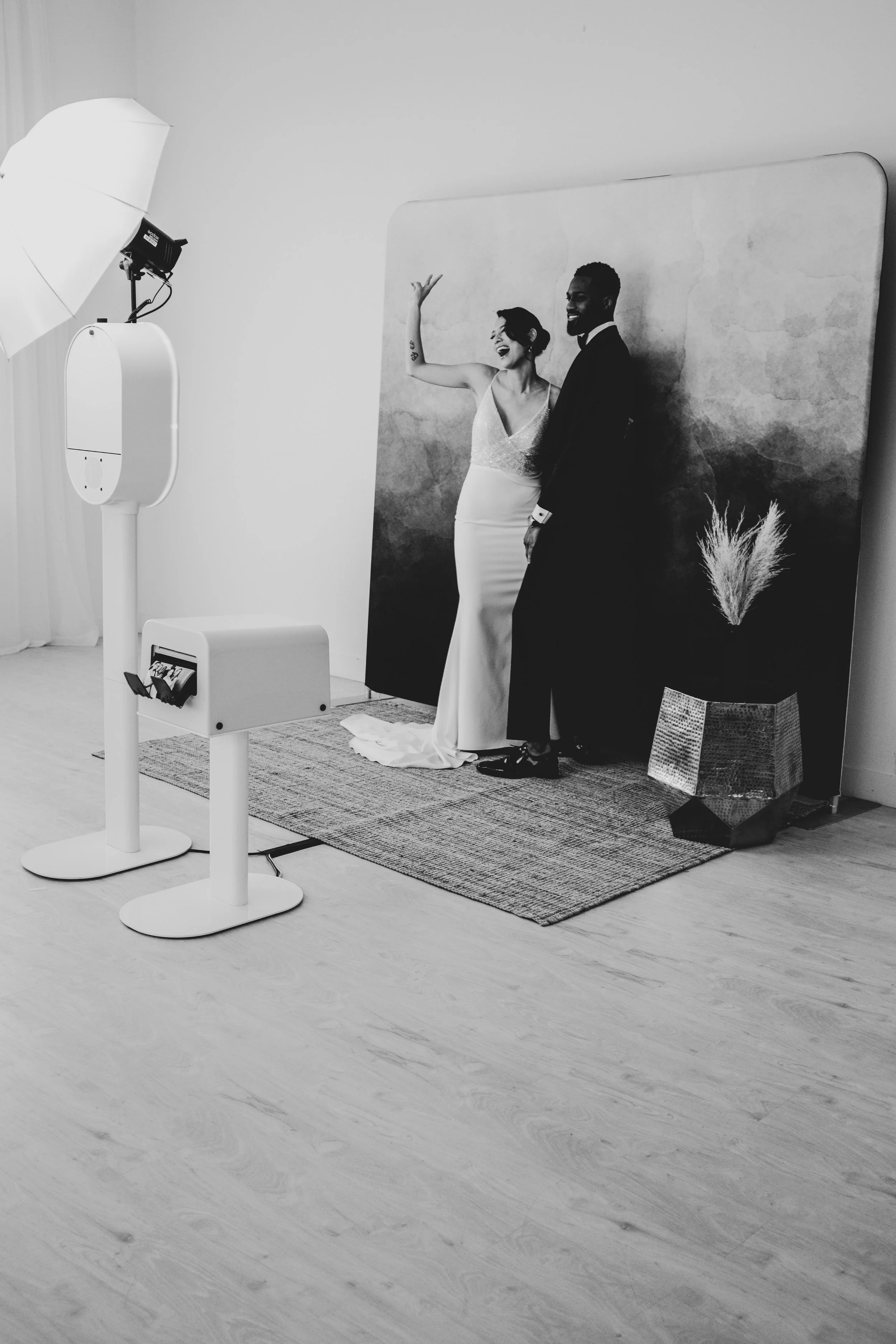 Black and white photo of a bride and groom posing for a wedding photo in front of a backdrop, with photo booth equipment nearby.