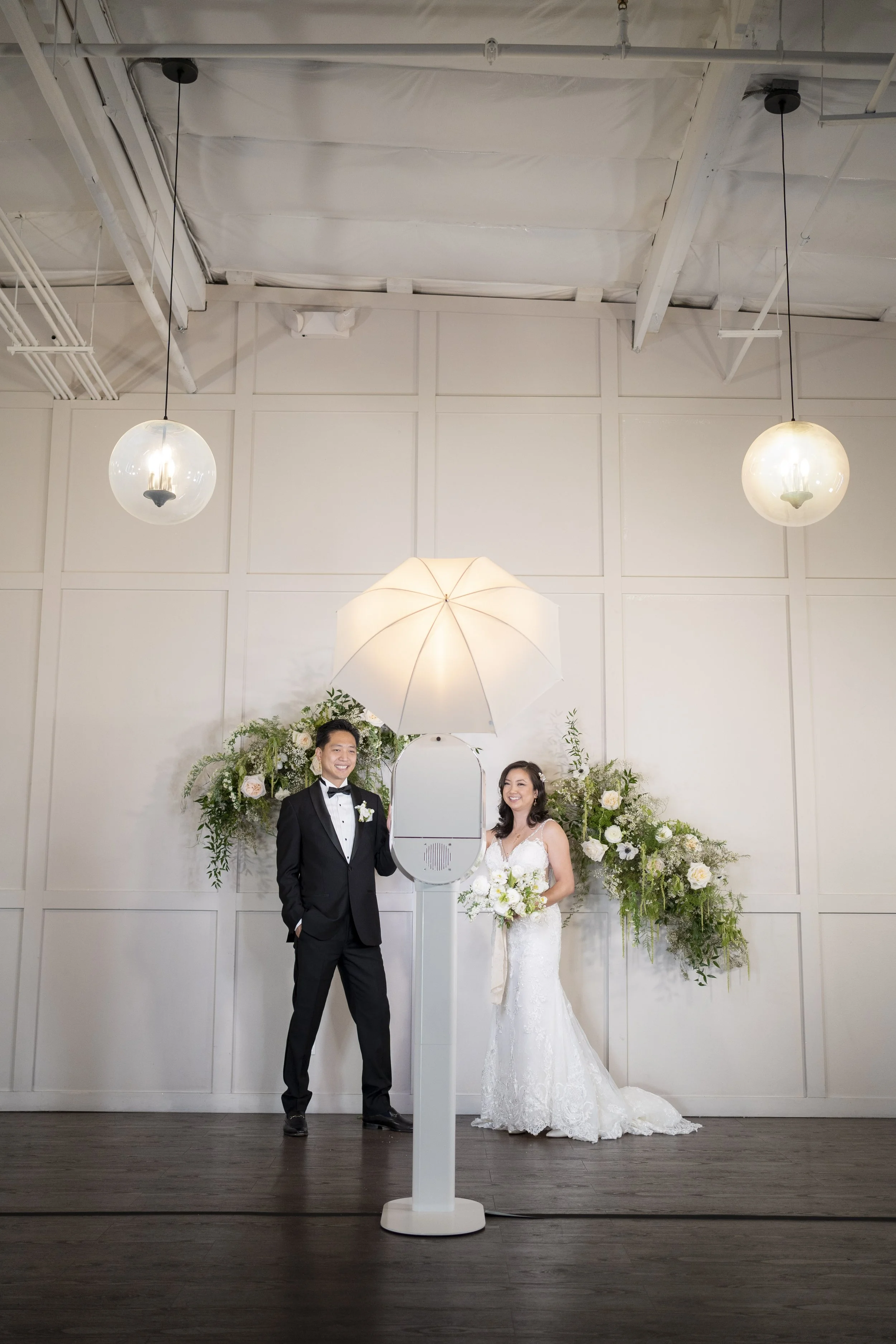 A bride and groom dressed in wedding attire celebrating, standing in front of a white wall with floral decorations, with a photo booth that has a large umbrella and two hanging lights overhead.