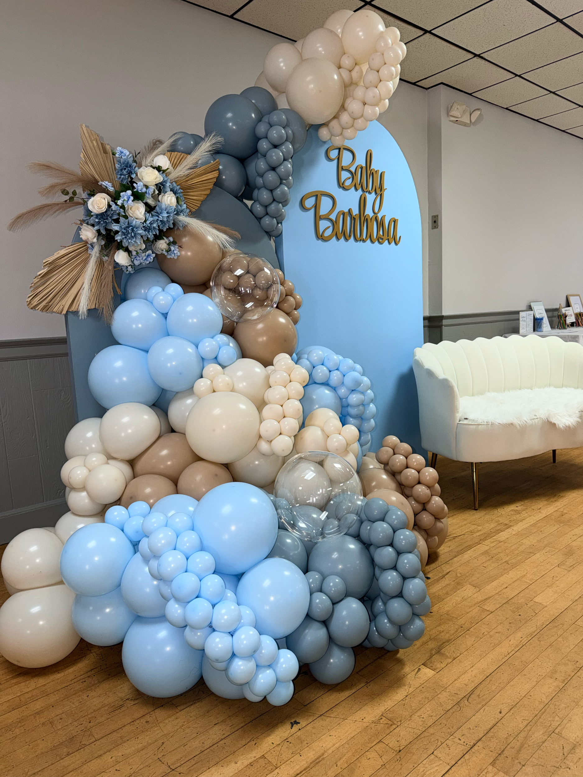 A decorative balloon arrangement with blue, beige, and white balloons, a flower bouquet with blue and white flowers and dried leaves, and a blue backdrop with the words 'Baby Barbsa' in gold lettering, next to a white upholstered chair.
