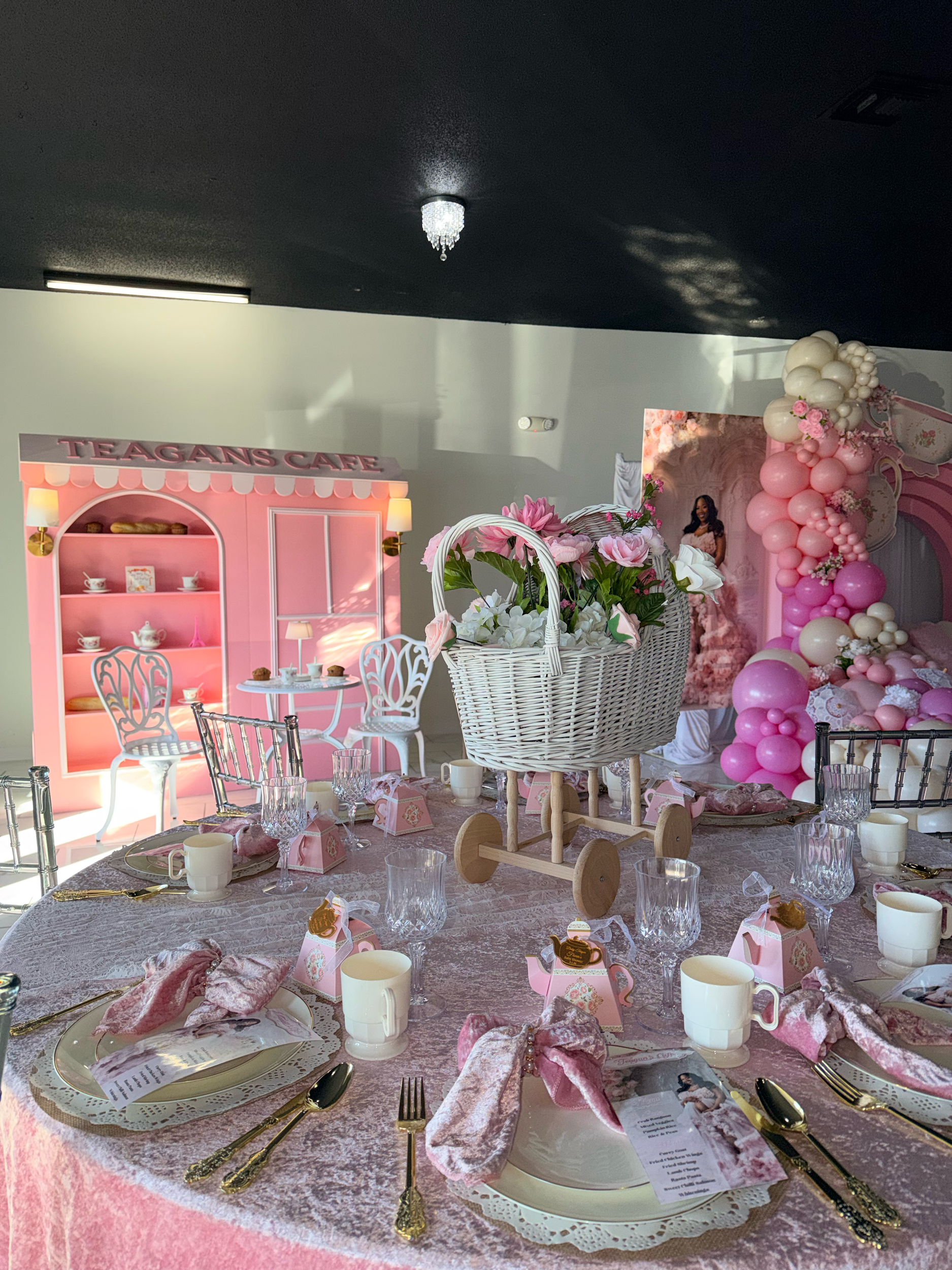 Decorative pink-themed table setting with tea cups, plates, napkins, and gold cutlery, featuring a pink teapot display, floral arrangements, and balloon arch with pink and white balloons.