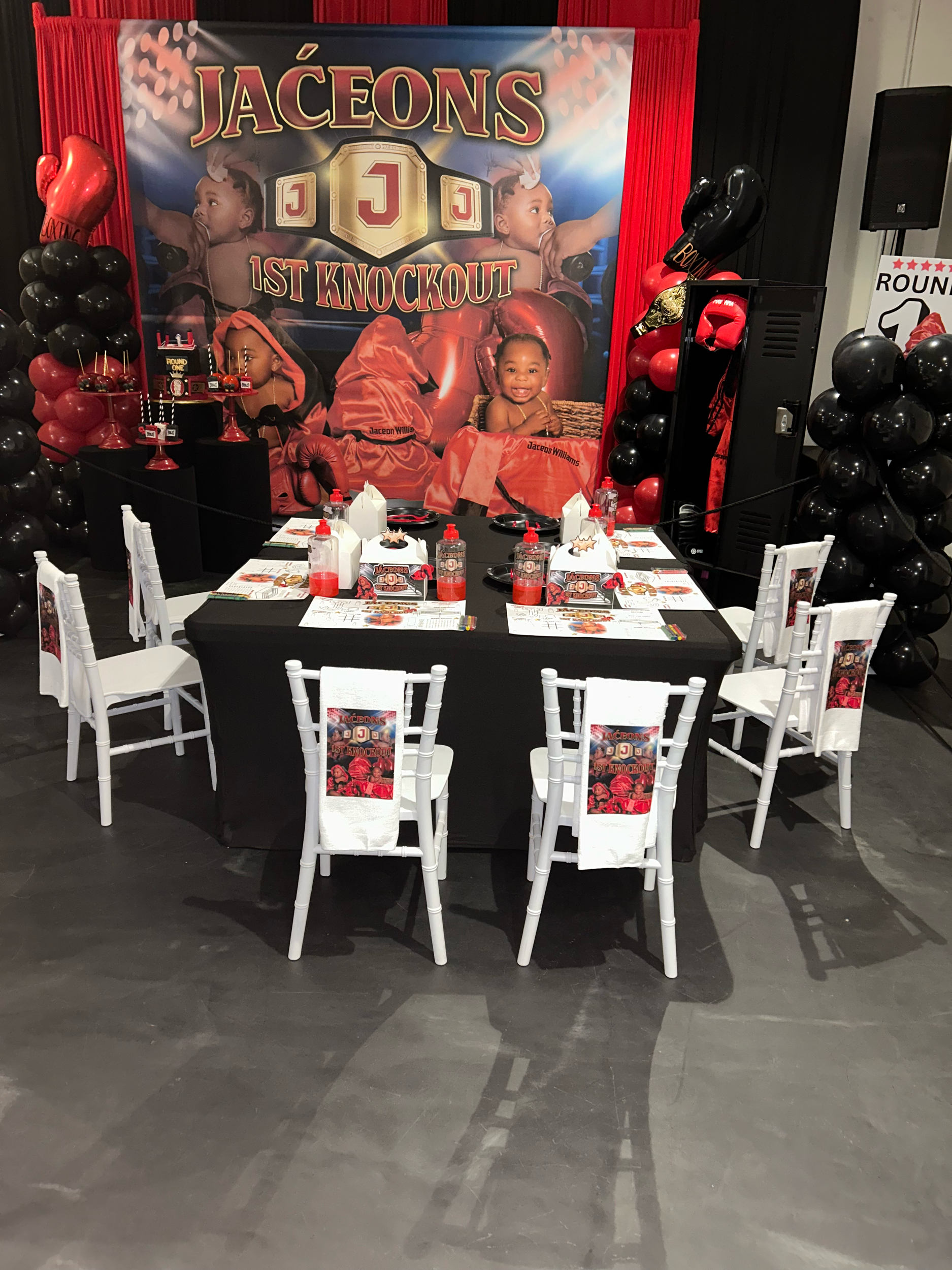 Party decoration with a large backdrop featuring a boxing theme, balloons, a table with party favors, and chairs for guests. The backdrop displays children in boxing gloves and costumes, with the text 'Jaceon 1st Knockout.'