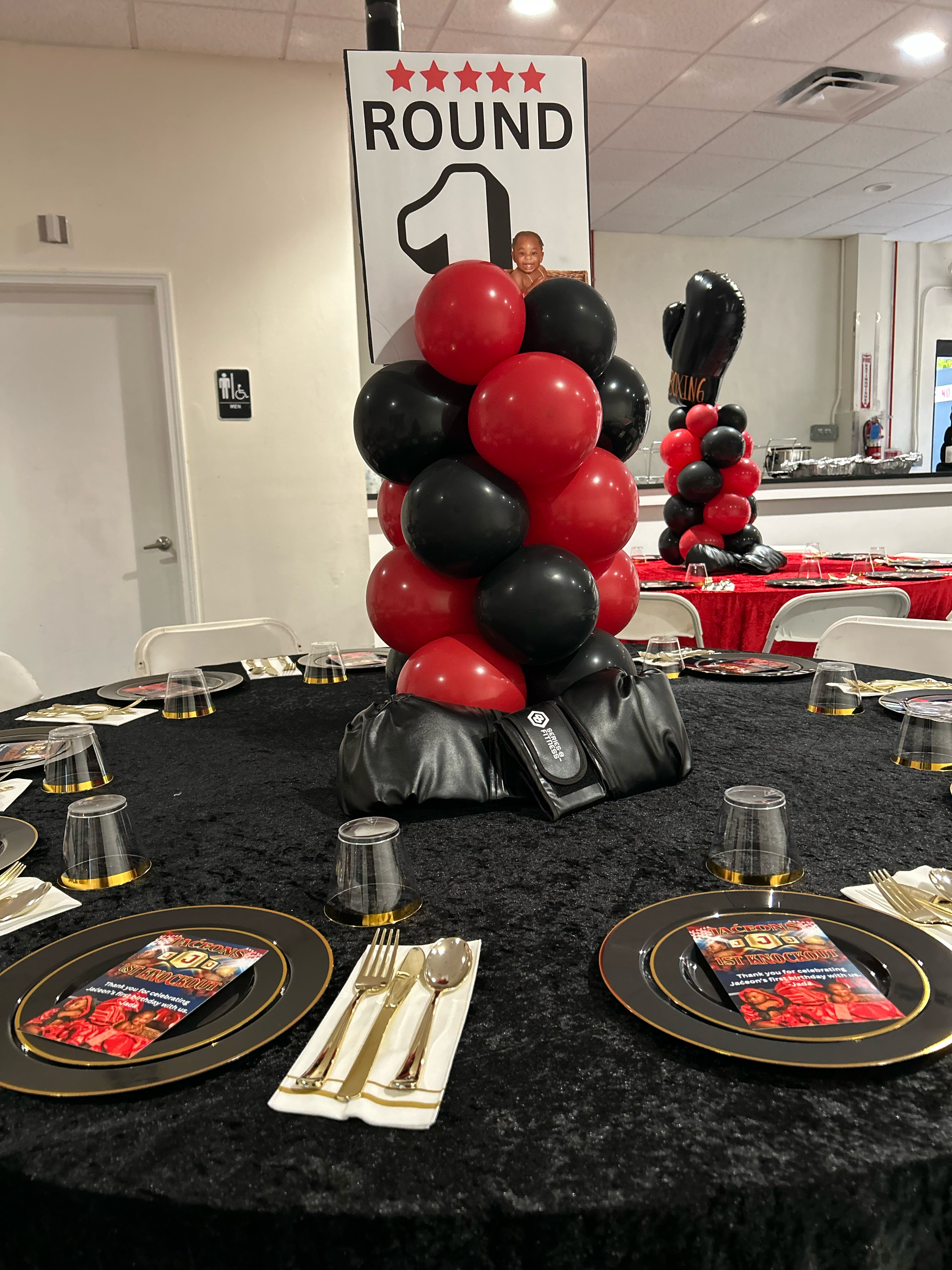 A table decorated for a birthday celebration with black, red, and gold-themed place settings, balloons, and a large sign indicating a fireworks-themed event. The sign features a large number '1' and a baby photo of a child. The table has plates with 