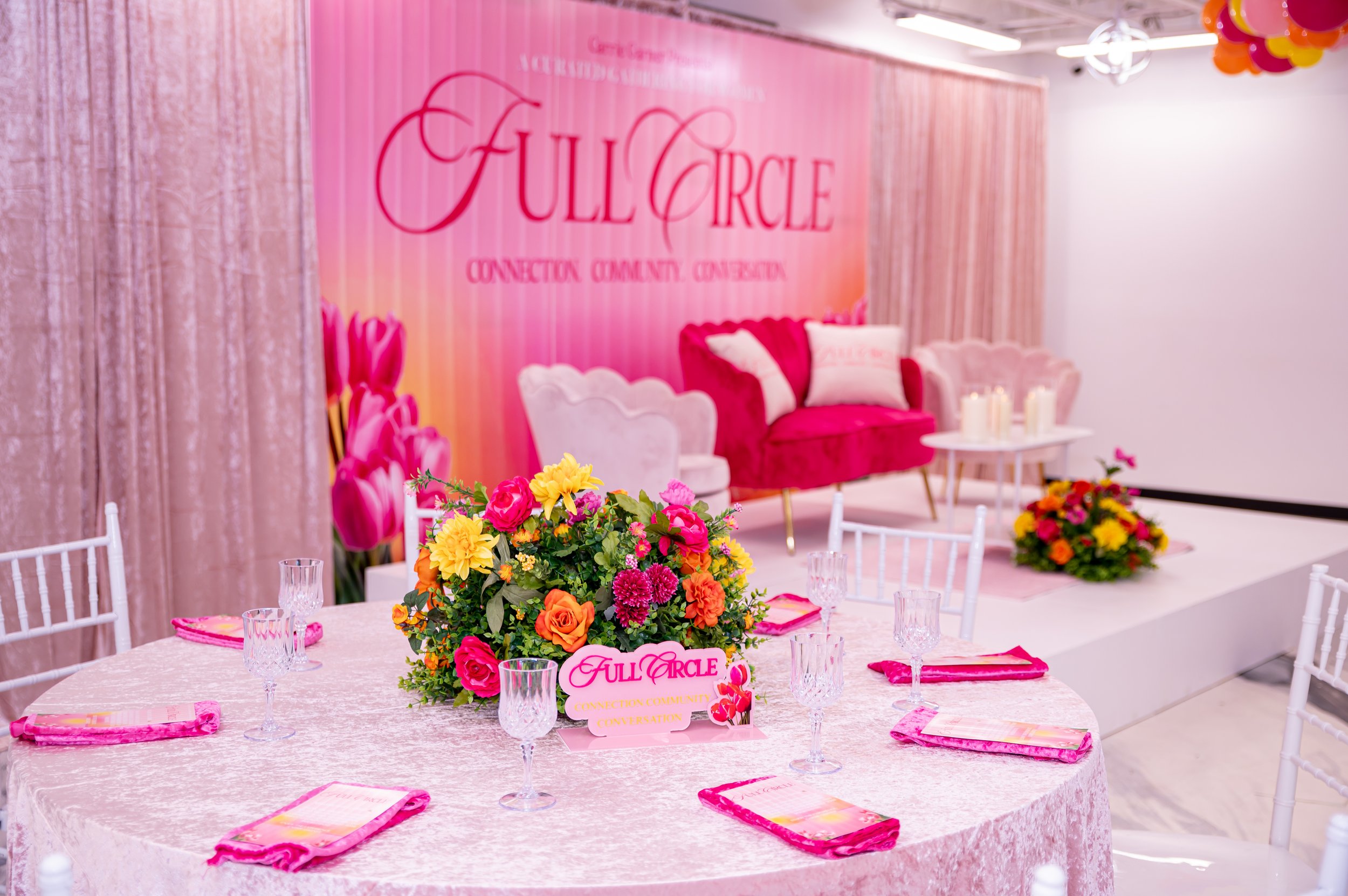 Decorated event space with a large pink floral centerpiece on a round table, pink napkins, and clear glassware. A pink and white sofa with pillows and a backdrop that reads "Full Circle Connection Community Conversation." Colorful balloons are also v