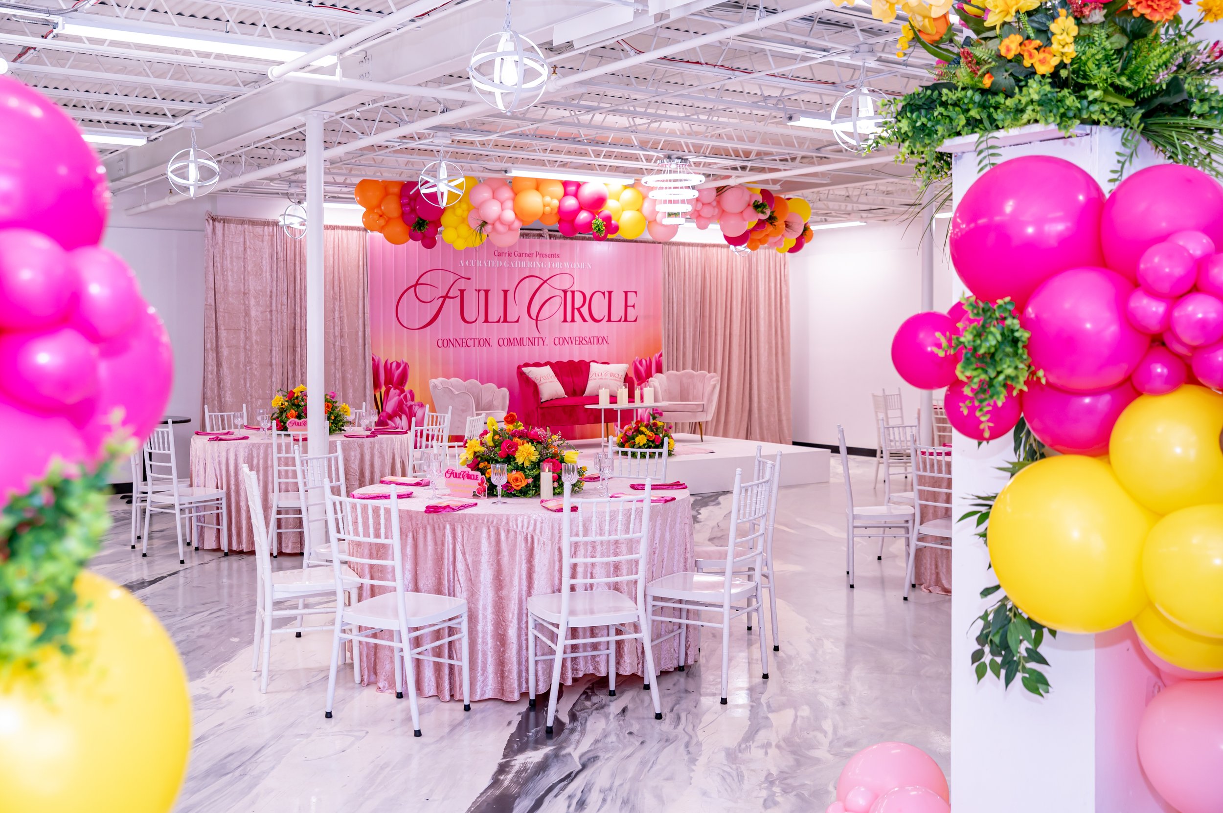 A decorated event space with pink, yellow, and orange balloons, floral arrangements, pink curtains, and a stage with a banner reading 'Full Circle,' featuring seating with pink and white decor for a celebration or gathering.