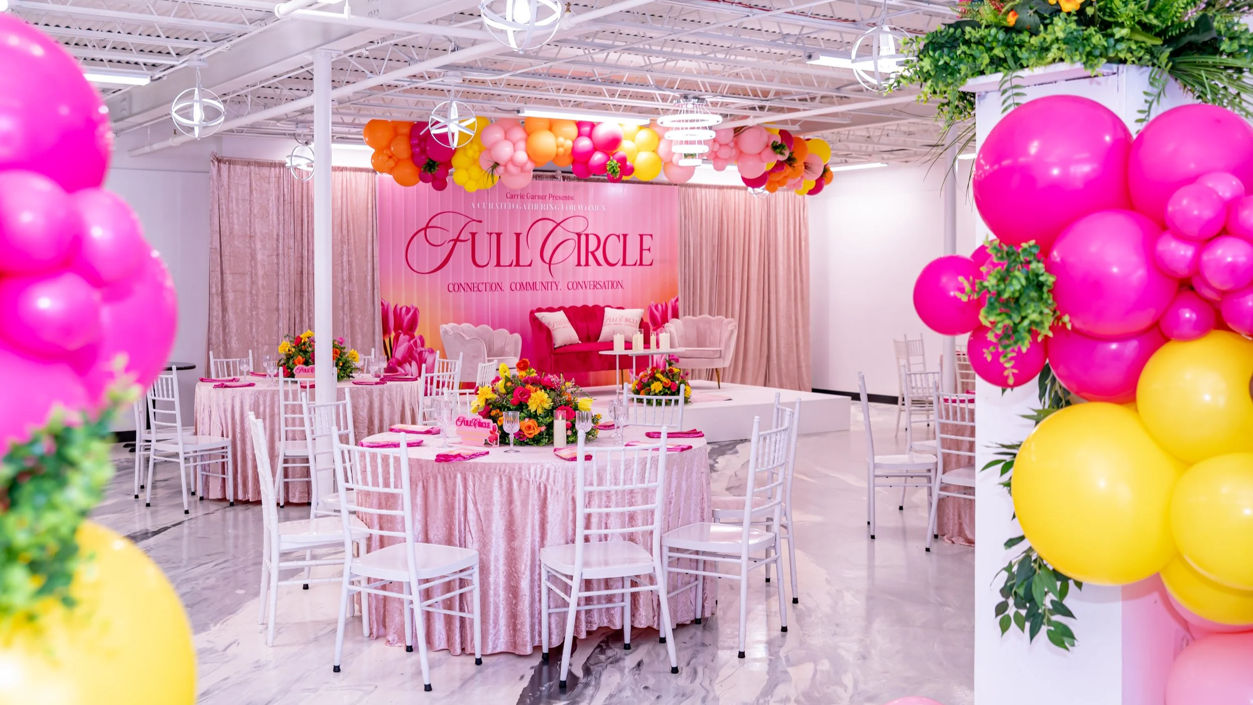 A decorated event space with pink and yellow balloons, pink curtains, a pink themed backdrop with the words 'Full Circle' and the phrases 'Connection. Community. Conversation.', round tables with pink tablecloths, floral centerpieces, and white chairs. The space appears to be prepared for a women’s gathering or celebration.