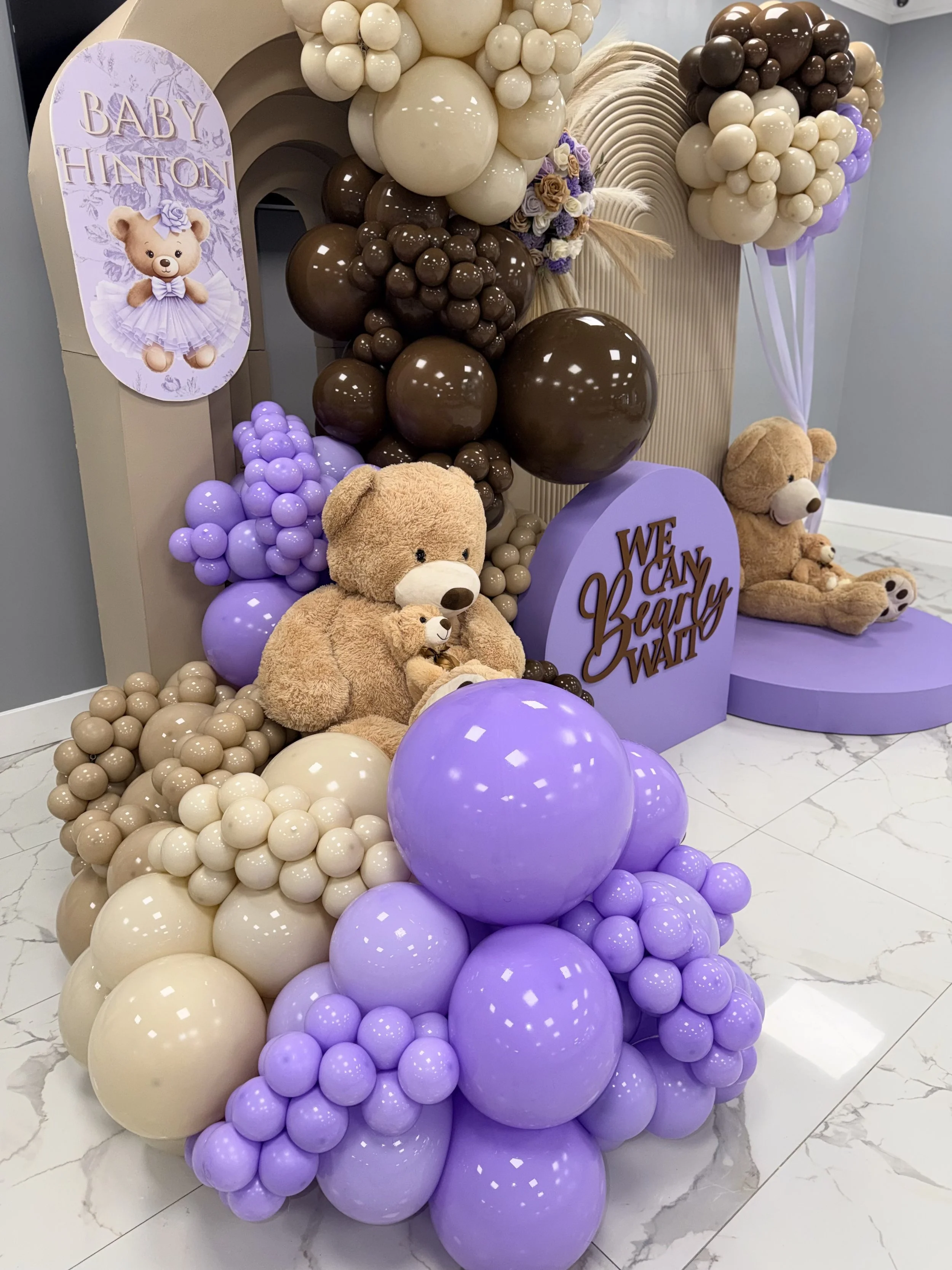 A baby shower balloon arrangement with cream, brown, and lavender balloons, teddy bears, and a sign that reads "Baby Hinton" and "We Can Beary Happy".