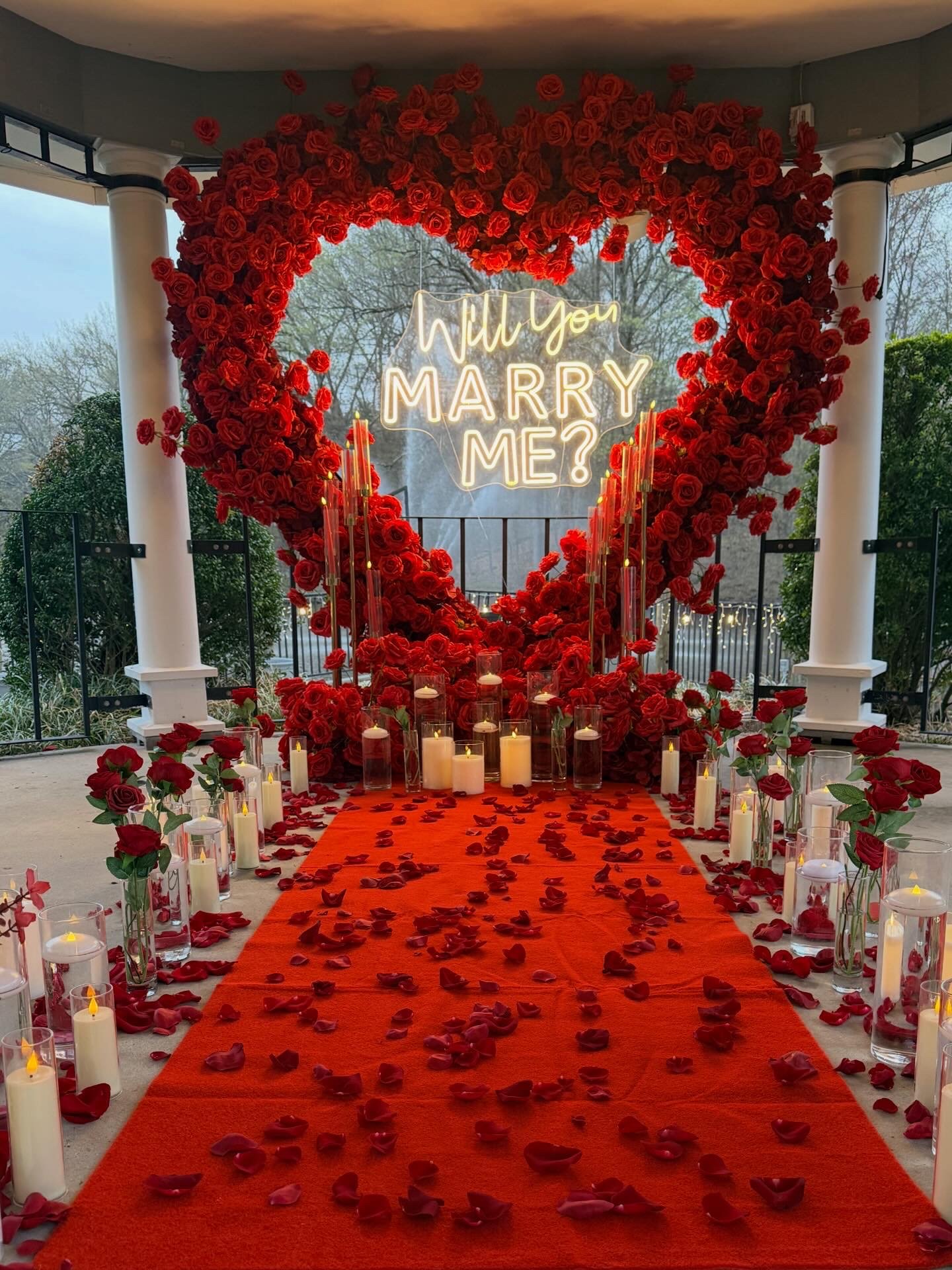 A love-themed wedding proposal setup with a large heart-shaped flower arrangement made of red roses, illuminated sign that says 'Will you marry me?', surrounded by candles, rose petals on an orange aisle runner, and outdoor scenery in the background.