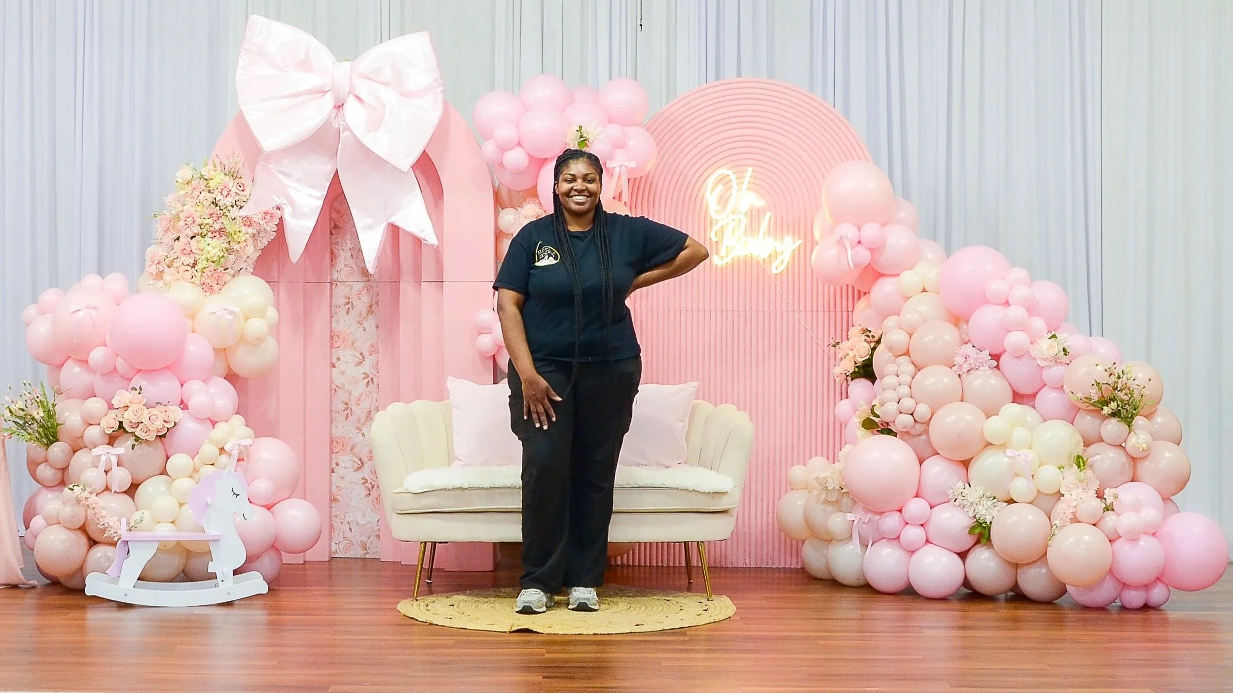 A woman standing in front of a pink-themed birthday decoration with balloons, flowers, a neon sign that reads 'Oh Baby,' and a white rocking horse.