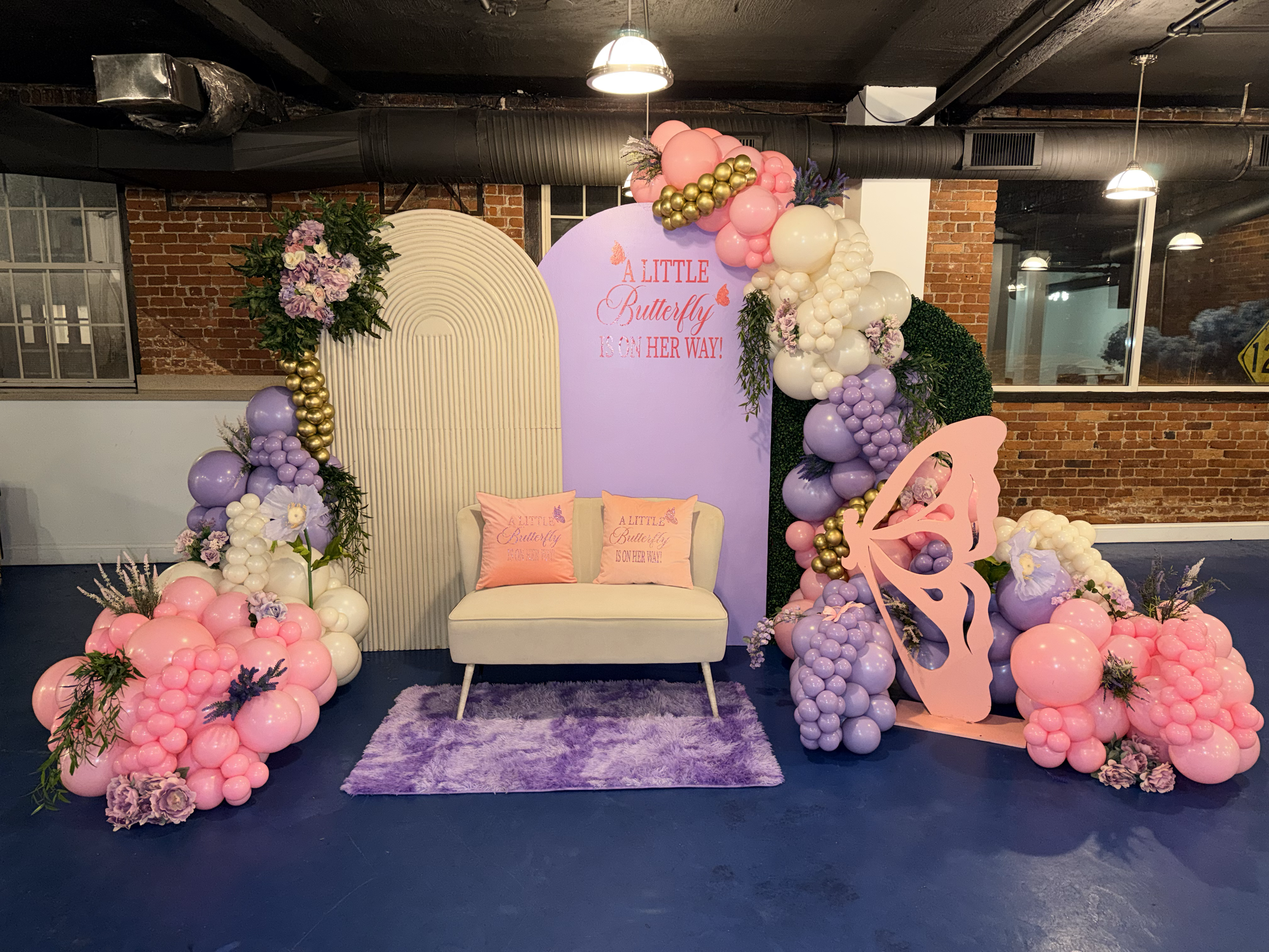 Decorative display with pink, purple, and white balloons arranged around a small white sofa with two pink pillows. Behind the sofa is a pink and purple backdrop with the phrase "A Little Butterfly Is On Her Way!" Fluffy purple rug on the floor, flora