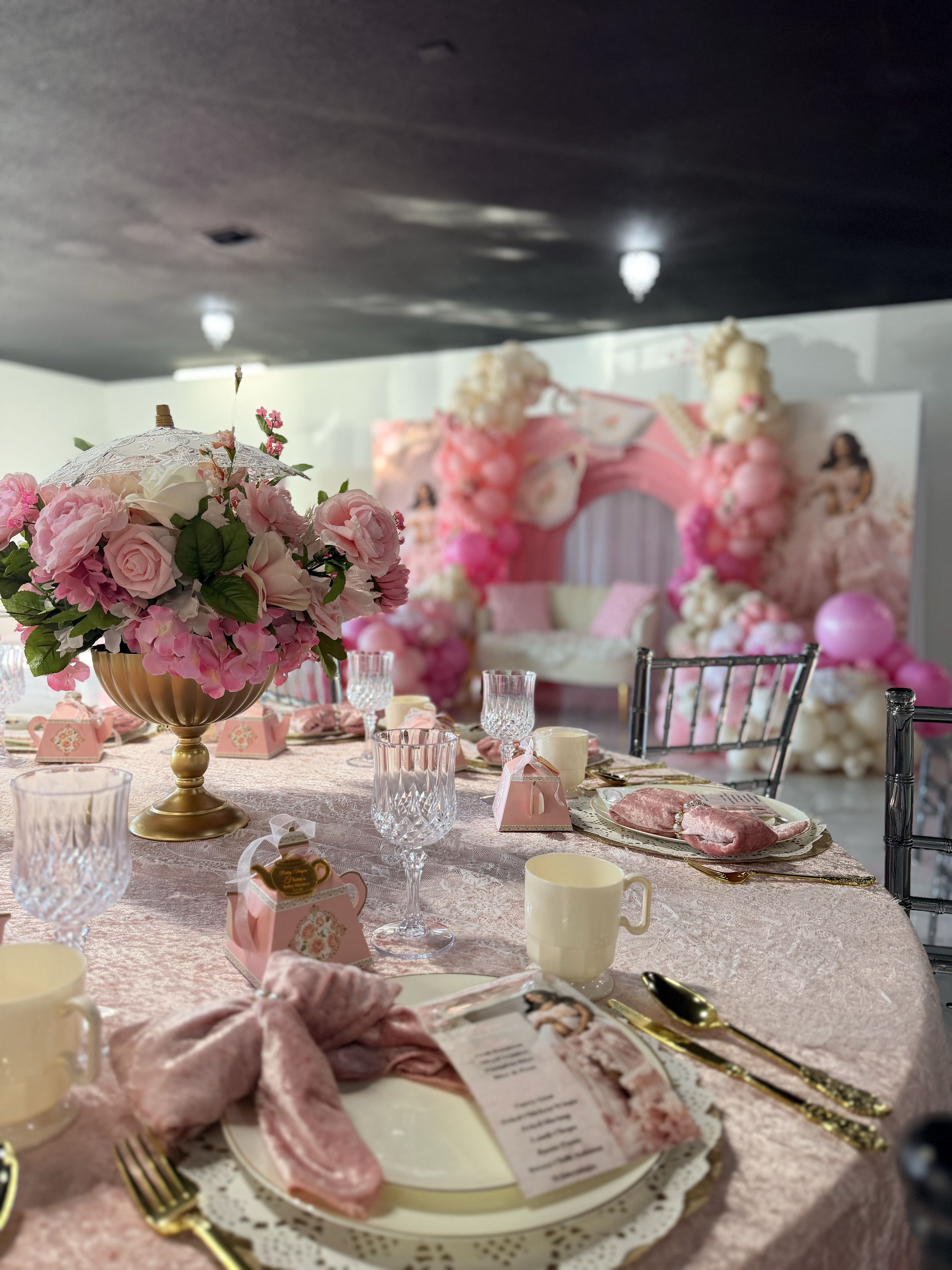 A pink-themed party table with a centerpiece of pink and white flowers, surrounded by glassware, gold utensils, and pink party favor boxes. In the background, a pink and white floral arch decorated with balloons and figures suggests a festive celebra