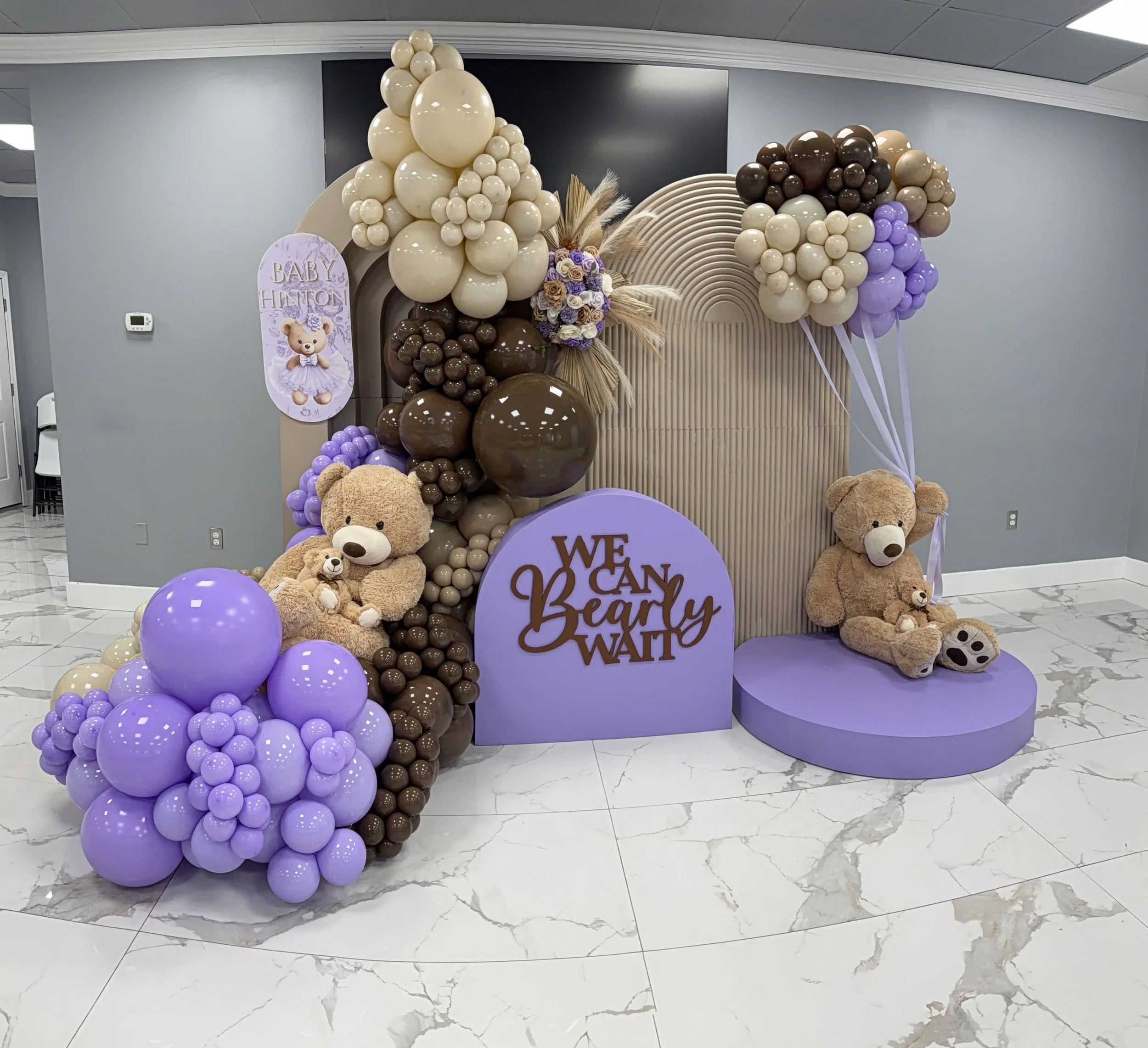 Decorative arrangement for a baby shower with balloons in shades of purple, beige, and brown, teddy bears, and a sign that reads 'Baby Henton, We Can't Be Bothered'.
