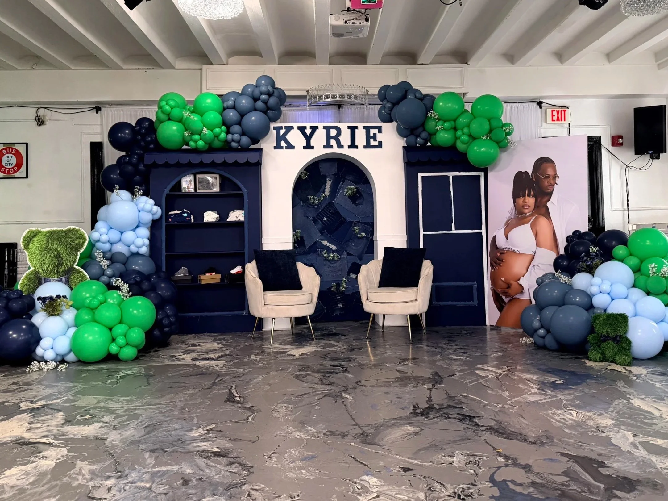 Decorative event backdrop featuring the name 'Kyrie' with green, black, gray, and white balloons arranged around a navy blue and white structure, with a large poster of a woman and baby, and two white mansions with black cushions in front, set on a marble floor.