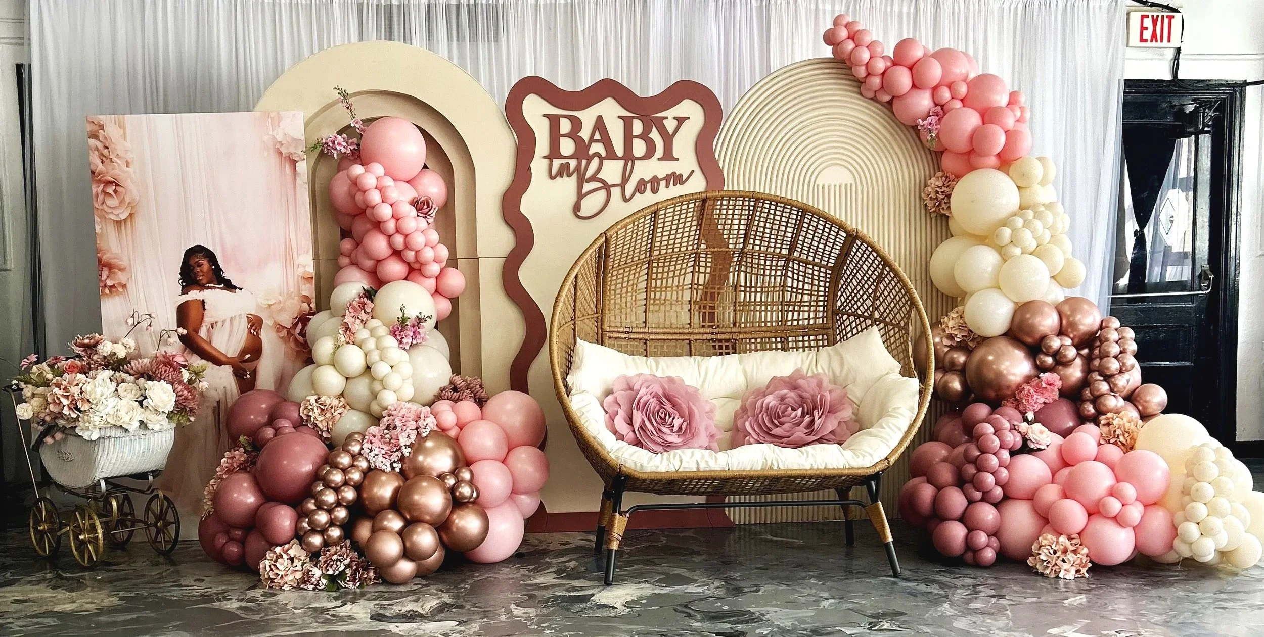 Decorative baby shower backdrop with balloons in pink, white, and rose gold, featuring a sign that says 'Baby Bloom,' a wicker seat with flower pillows, a 'New Arrival' photo display, and floral arrangements.