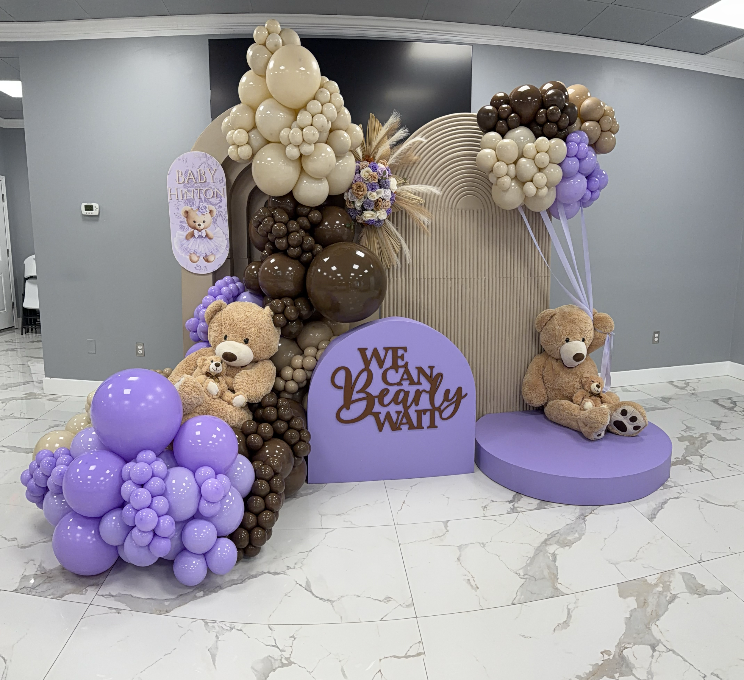Balloon arch in colors of cream, brown, and purple, decorated with teddy bears and a floral arrangement, with a sign that says "Baby Hinton" and a purple circle sign that says "We Can Bearly Wait" for a baby shower celebration.