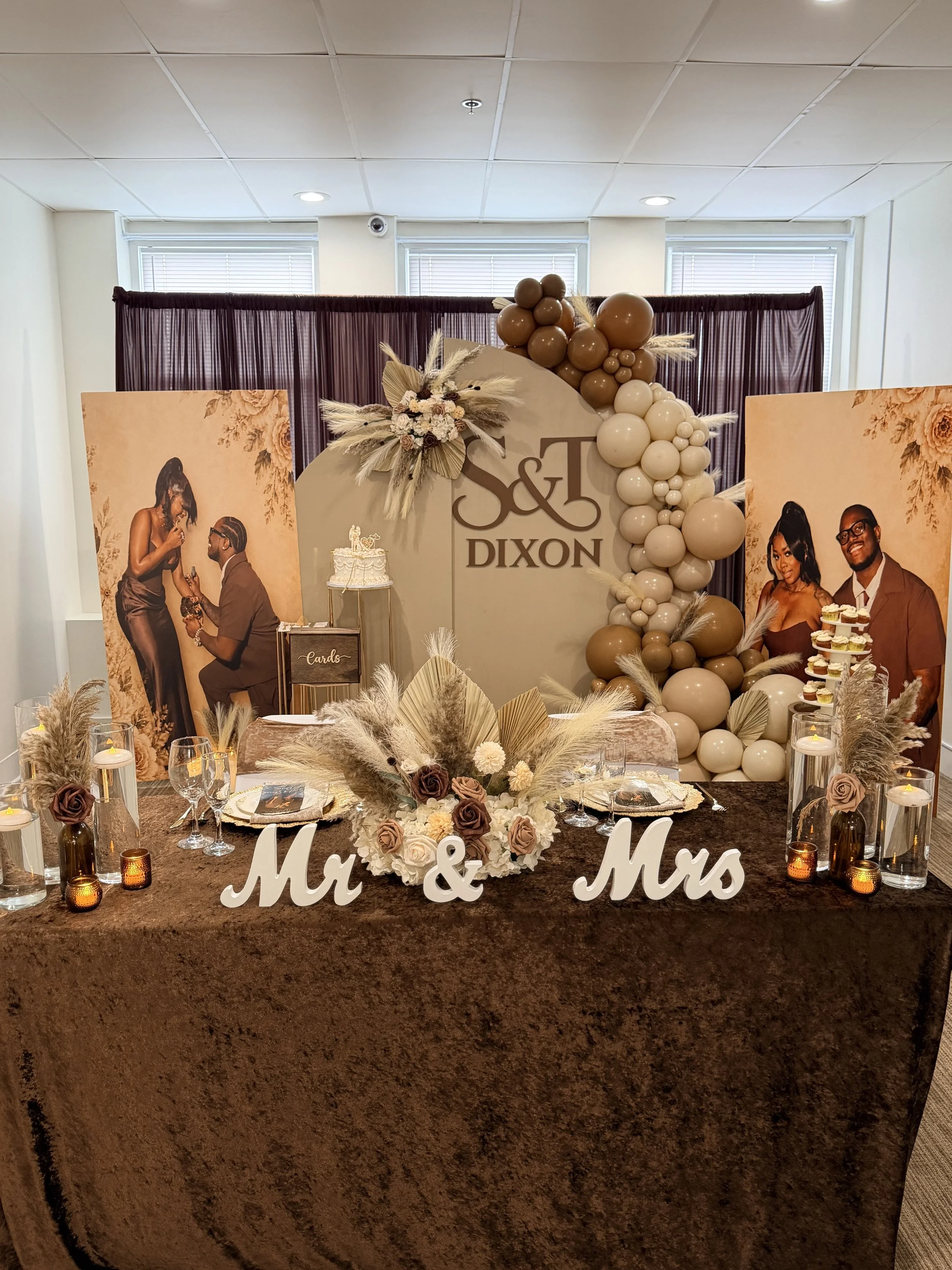 A wedding reception display with a large round backdrop decorated with balloons in beige, white, and brown shades, and the initials 'S & T' with the last name Dixon. There are two vertical posters showing the couple, a cake on a stand, and a table with floral arrangements, candles, and the sign 'Mr. & Mrs.' in front.