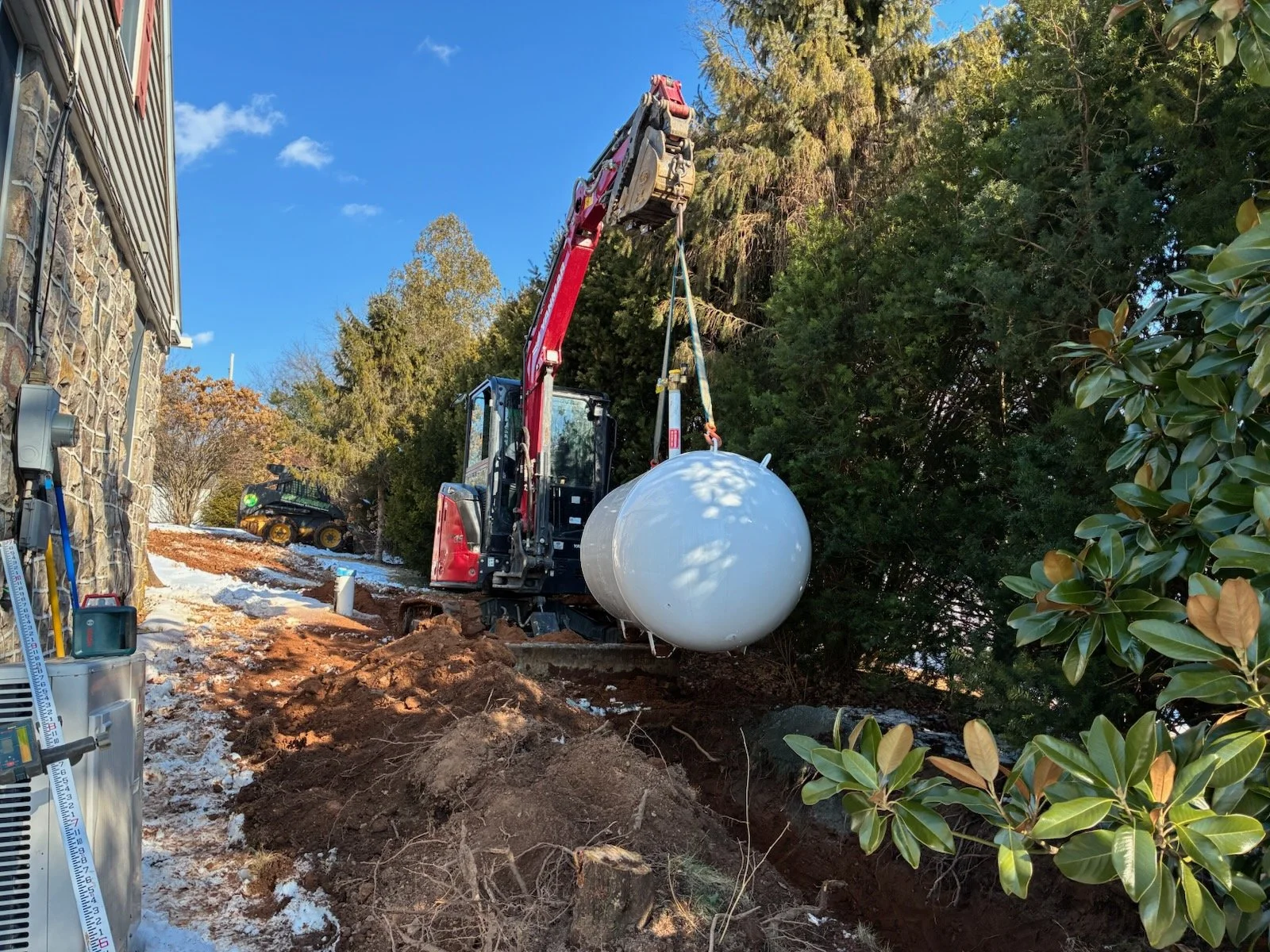 Construction site with a red and black excavator lifting a large white spherical tank. The site is next to a stone building and surrounded by trees, with some snow and dirt on the ground.