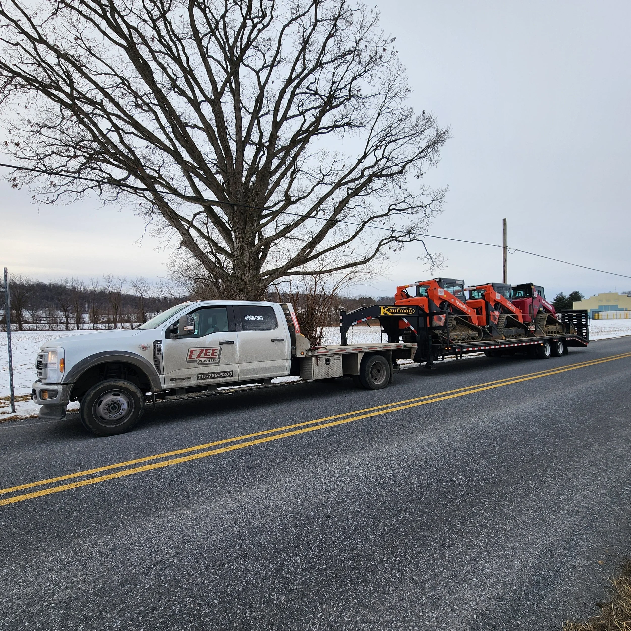 A flatbed truck carrying two orange construction vehicles parked on a road with a leafless tree in the background and snow on the ground.