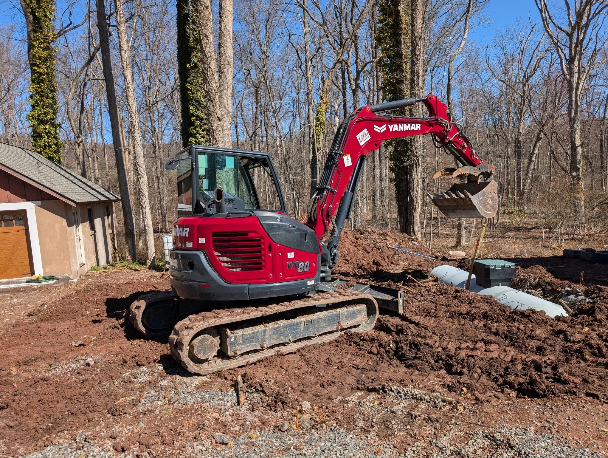 A red Yanmar mini excavator with tracked wheels working on outdoor construction, digging into soil near a house with a brown roof, surrounded by leafless trees on a clear day.