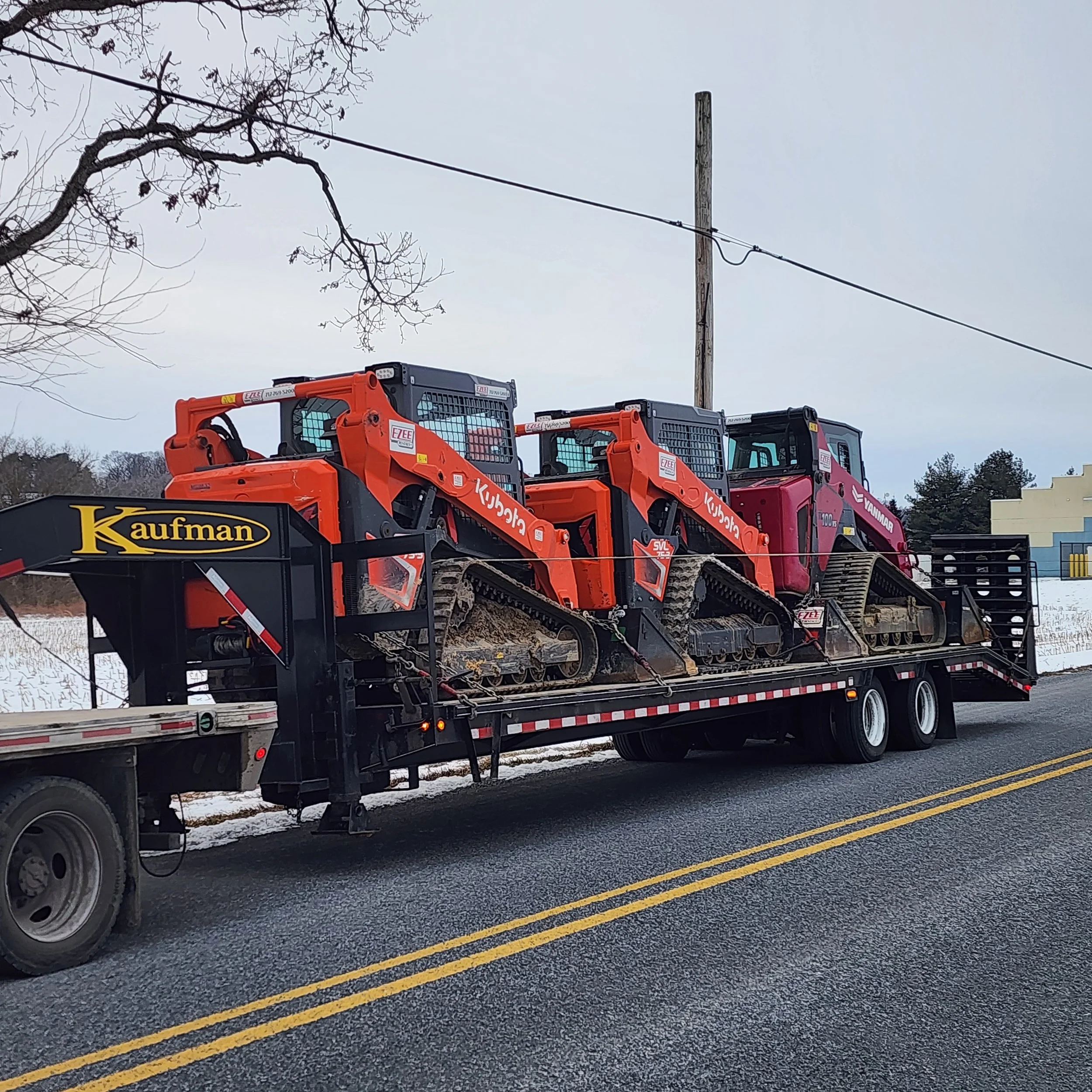 Two small orange tracked loaders and one larger pink tracked loader loaded on a flatbed trailer on a road with snow on the ground, an overhead utility line, and leafless trees.