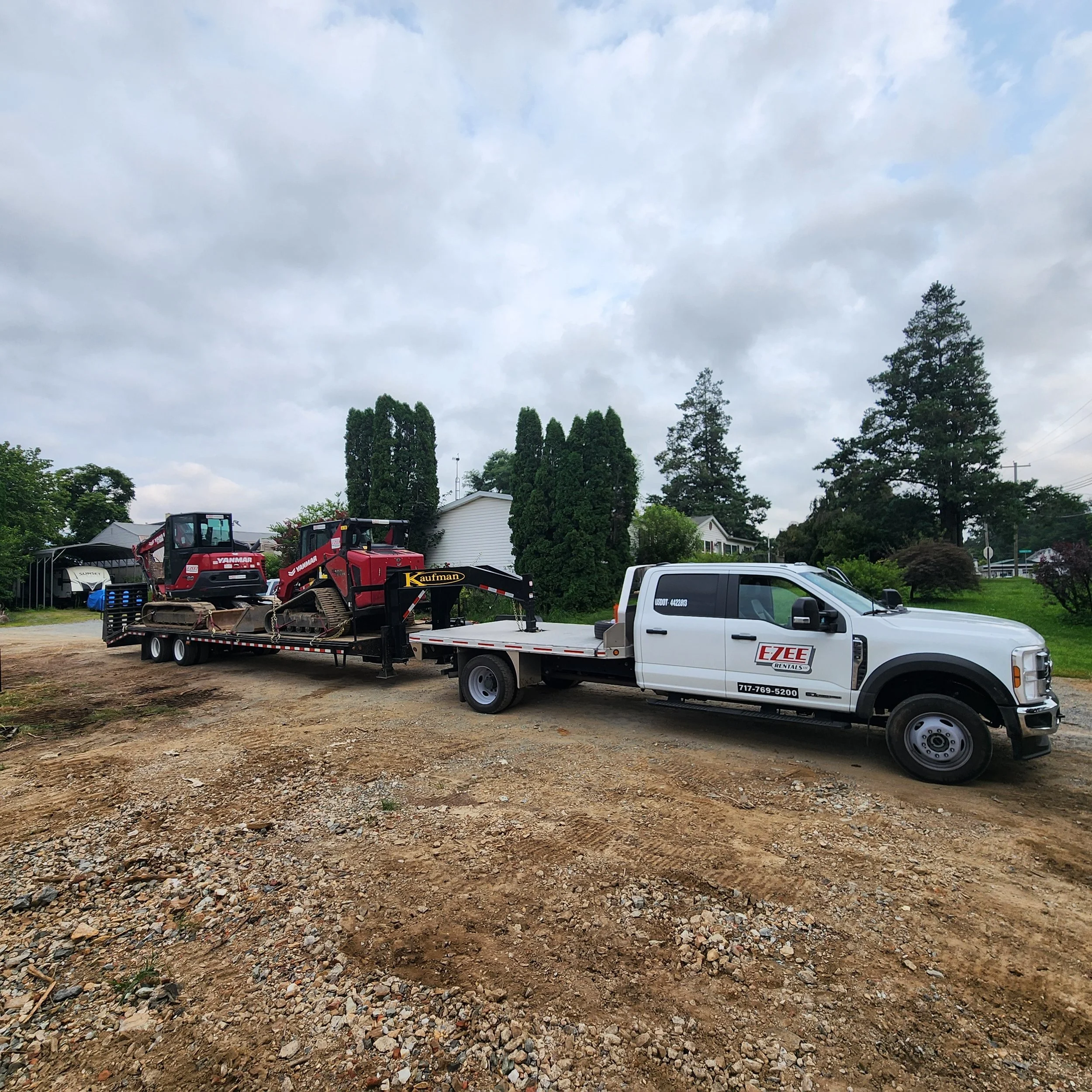 A white flatbed truck with EZEE rentals branding transporting two pieces of heavy equipment, a red excavator and a red tracked loader, on a dirt lot with green trees and cloudy sky in the background.