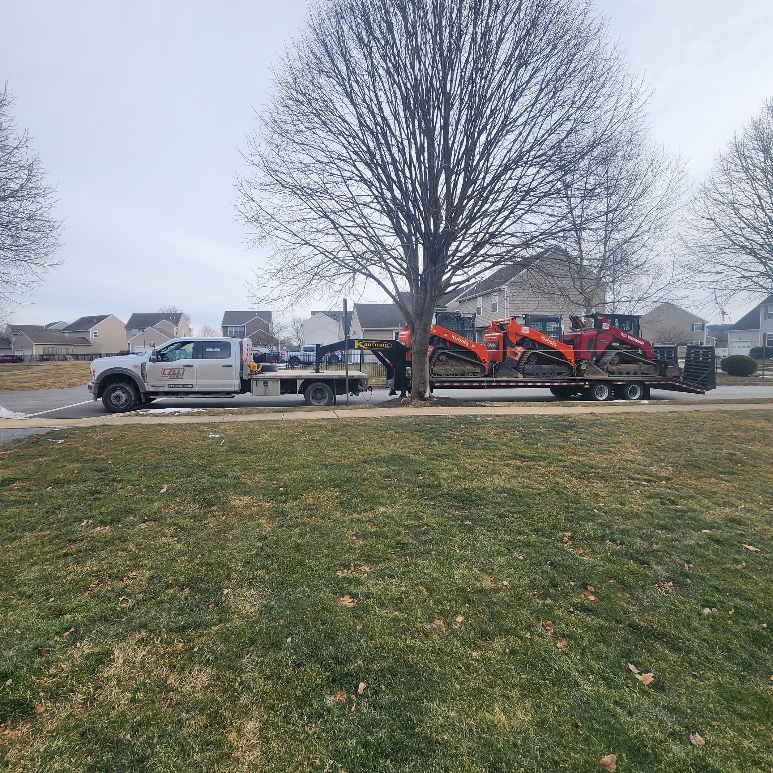 A pickup truck with a flatbed trailer carrying three colored track skid-steers is parked beside a leafless tree on a suburban street.