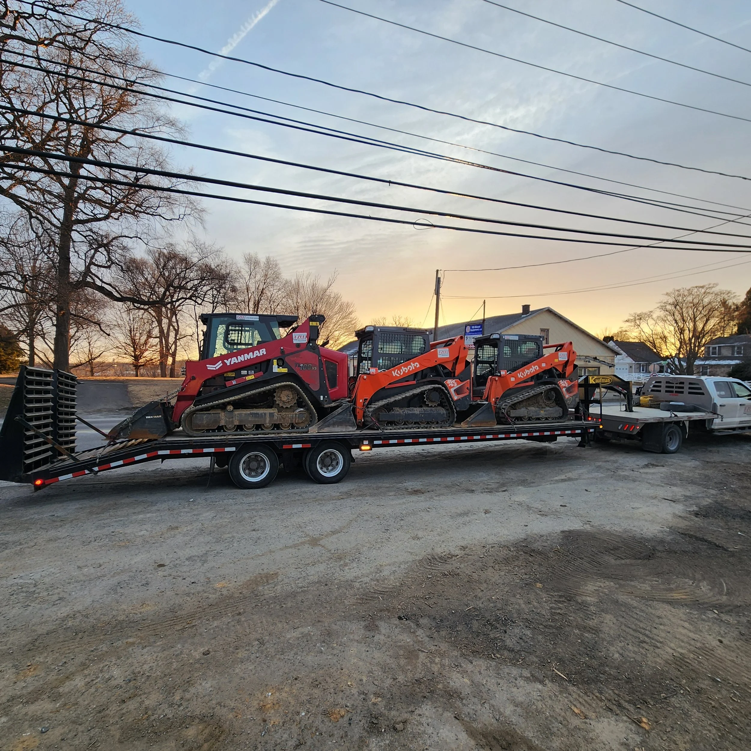 A flatbed trailer transporting three small compact loaders, with two orange Kubota loaders and one red Yanmar loader, parked on a dirt lot during sunset with trees, power lines, and residential houses in the background.