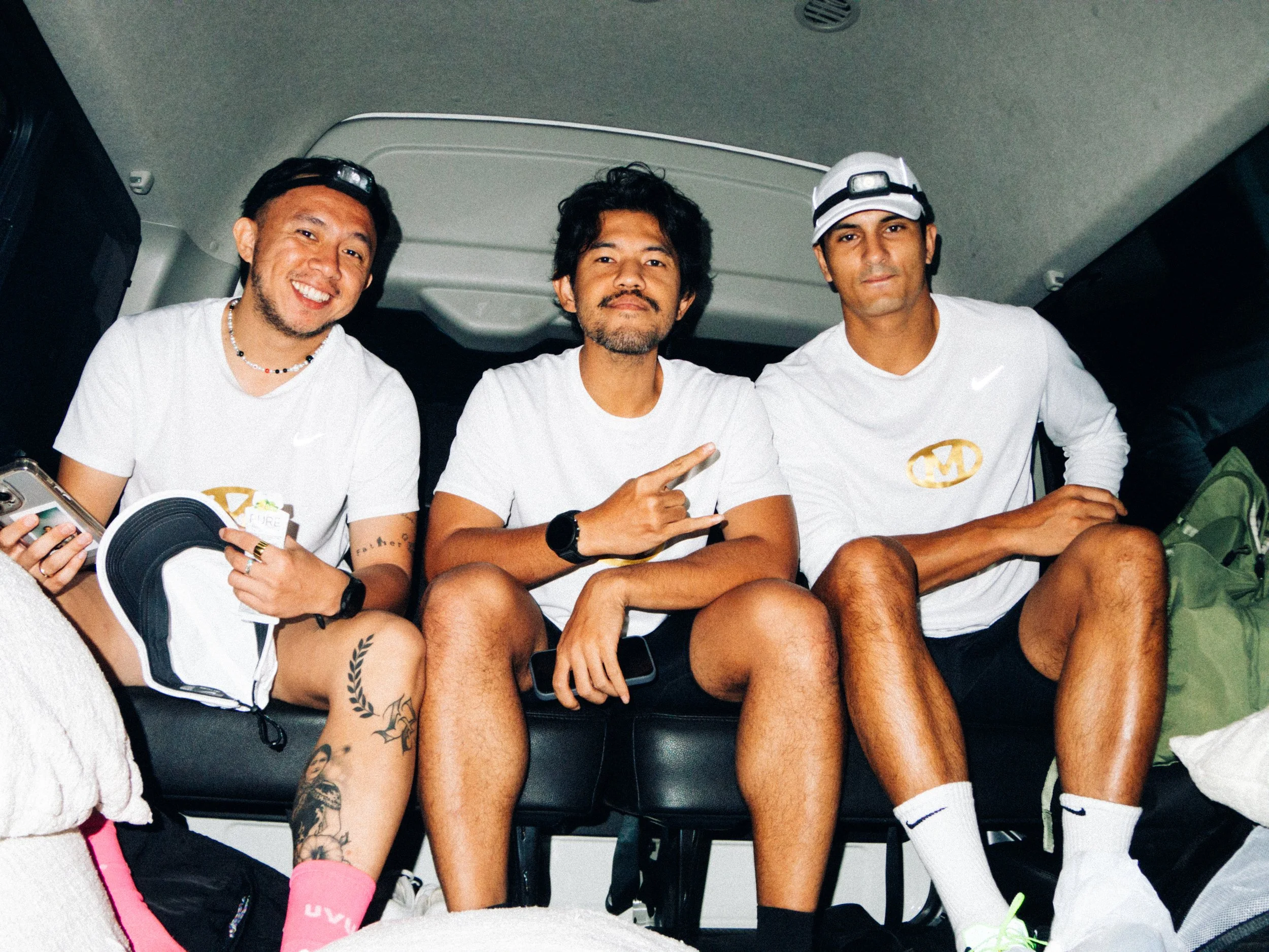 Three men sitting inside a vehicle, smiling at the camera. All wear white shirts, with the man on the right also wearing a cap and white socks, and the man on the left holding a soccer shoe. The man in the middle is making a peace sign. They are casu