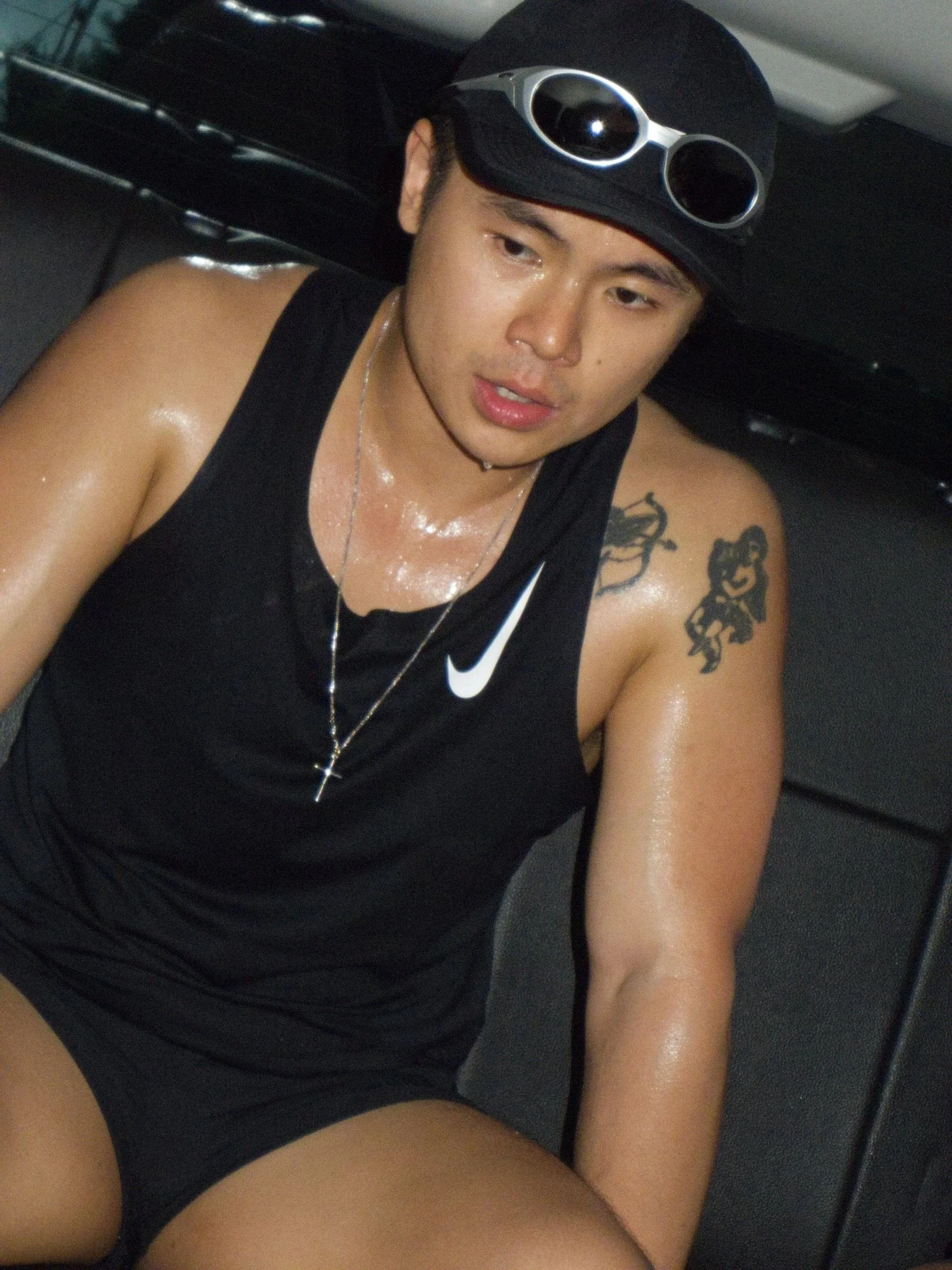 A young man wearing a black tank top, cap, and sunglasses, sweating, sitting on a black leather seat.