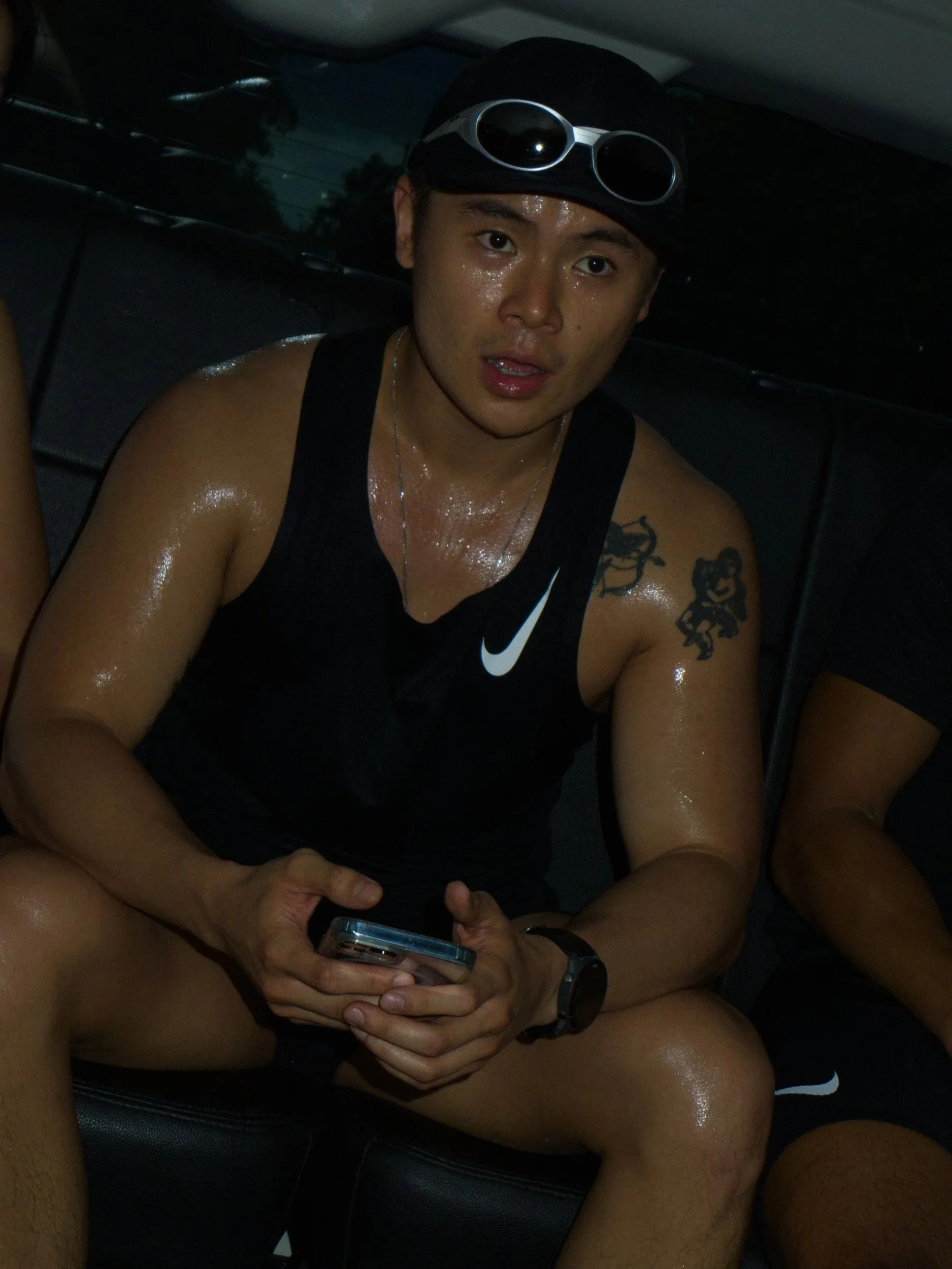 A young man with sunglasses on his head, wearing a black Nike tank top, sitting in a car with a wet, sweaty appearance, holding a smartphone in his hands.