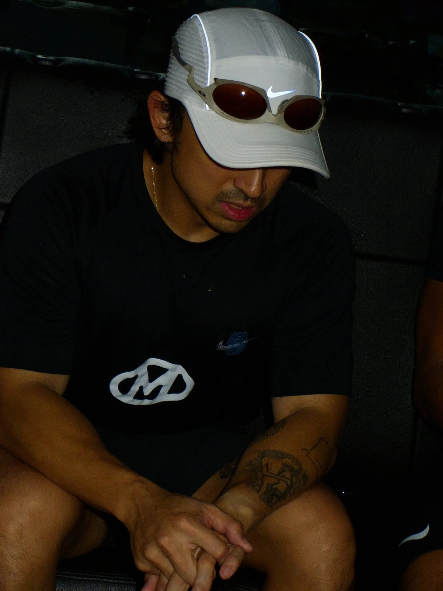 A young man wearing a white Nike baseball cap with sunglasses on top, a black t-shirt with designs, and tattoos on his arms, sitting with his hands clasped and looking down.