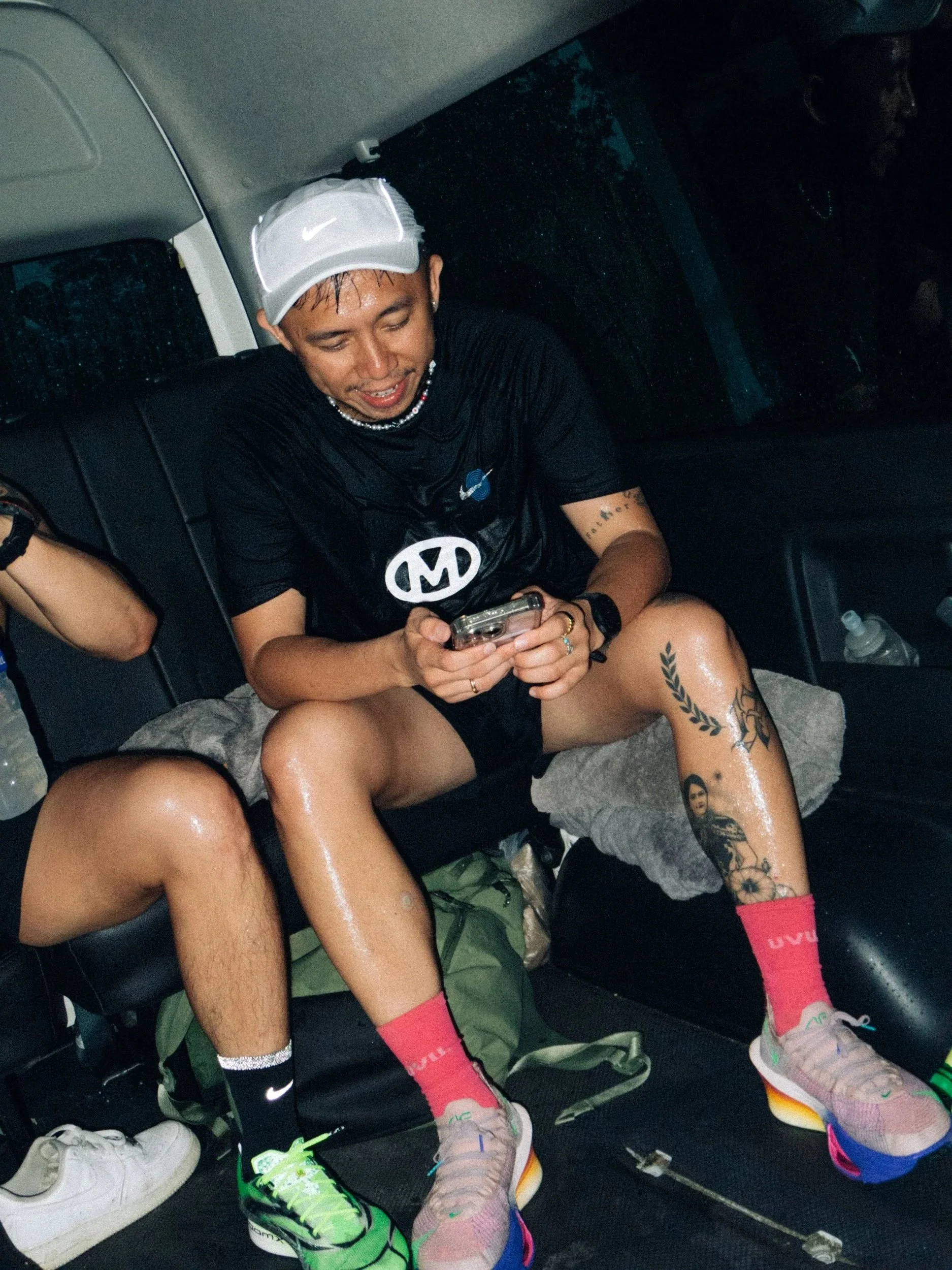 A man sitting in a vehicle, wearing sports shoes, colorful socks, a black T-shirt, a white hat, and with tattoos on his legs. He appears to be looking at his phone.