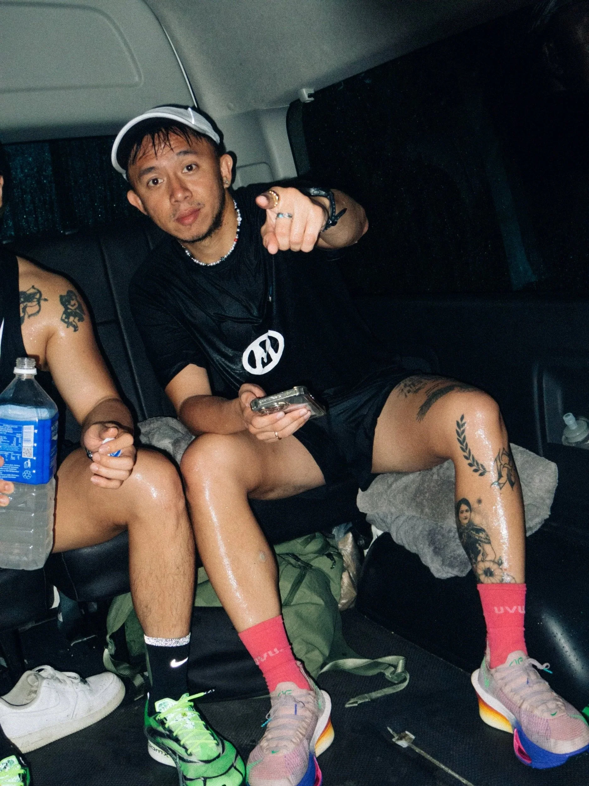 A man sitting in a vehicle with wet hair, wearing a black t-shirt with a peace symbol, red shorts, and colorful running shoes, pointing at the camera.