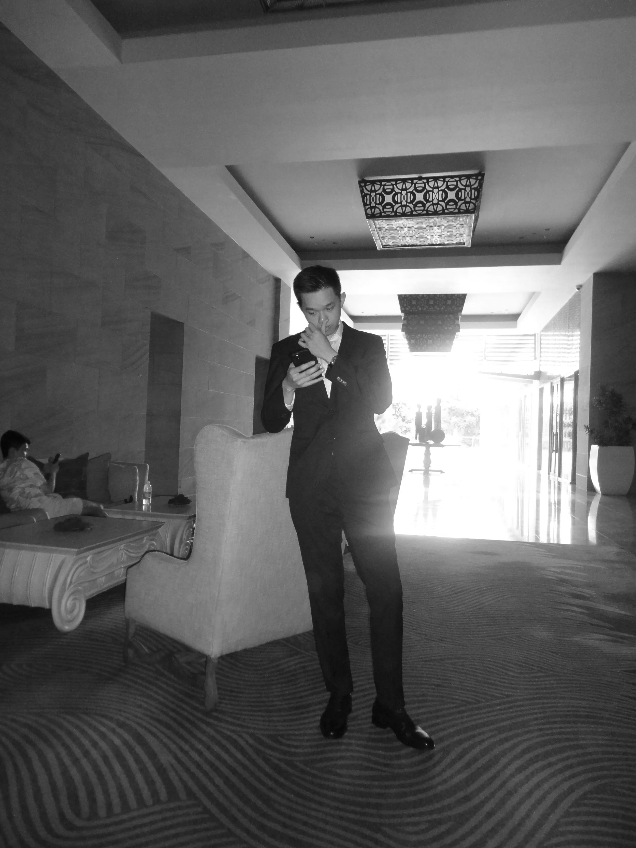 A man in a suit standing in a hotel lobby, looking at his phone, with a woman sitting on a nearby sofa in the background.