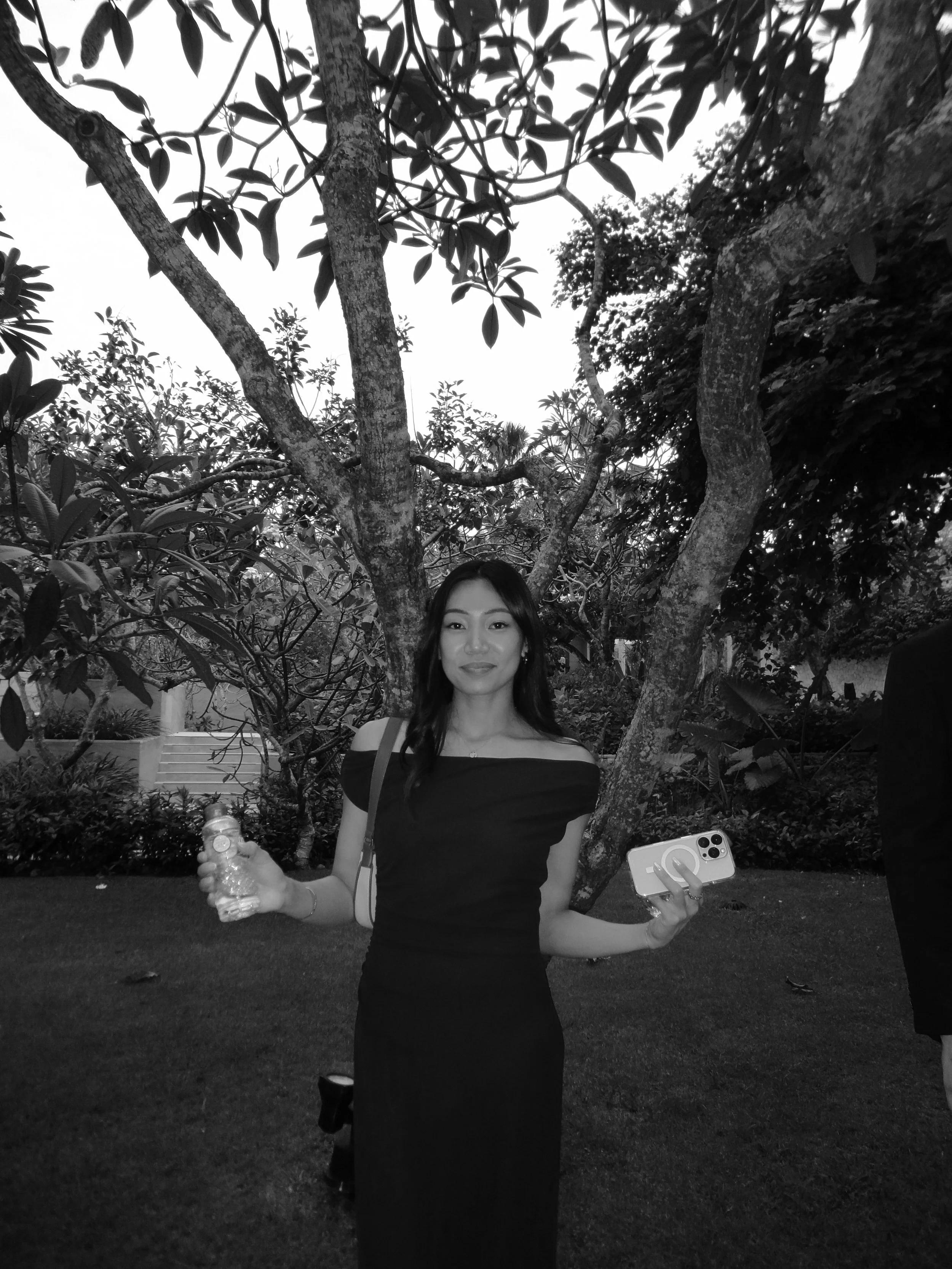 A woman in an off-shoulder dress standing outdoors in front of trees, holding a water bottle in one hand and a smartphone in the other.
