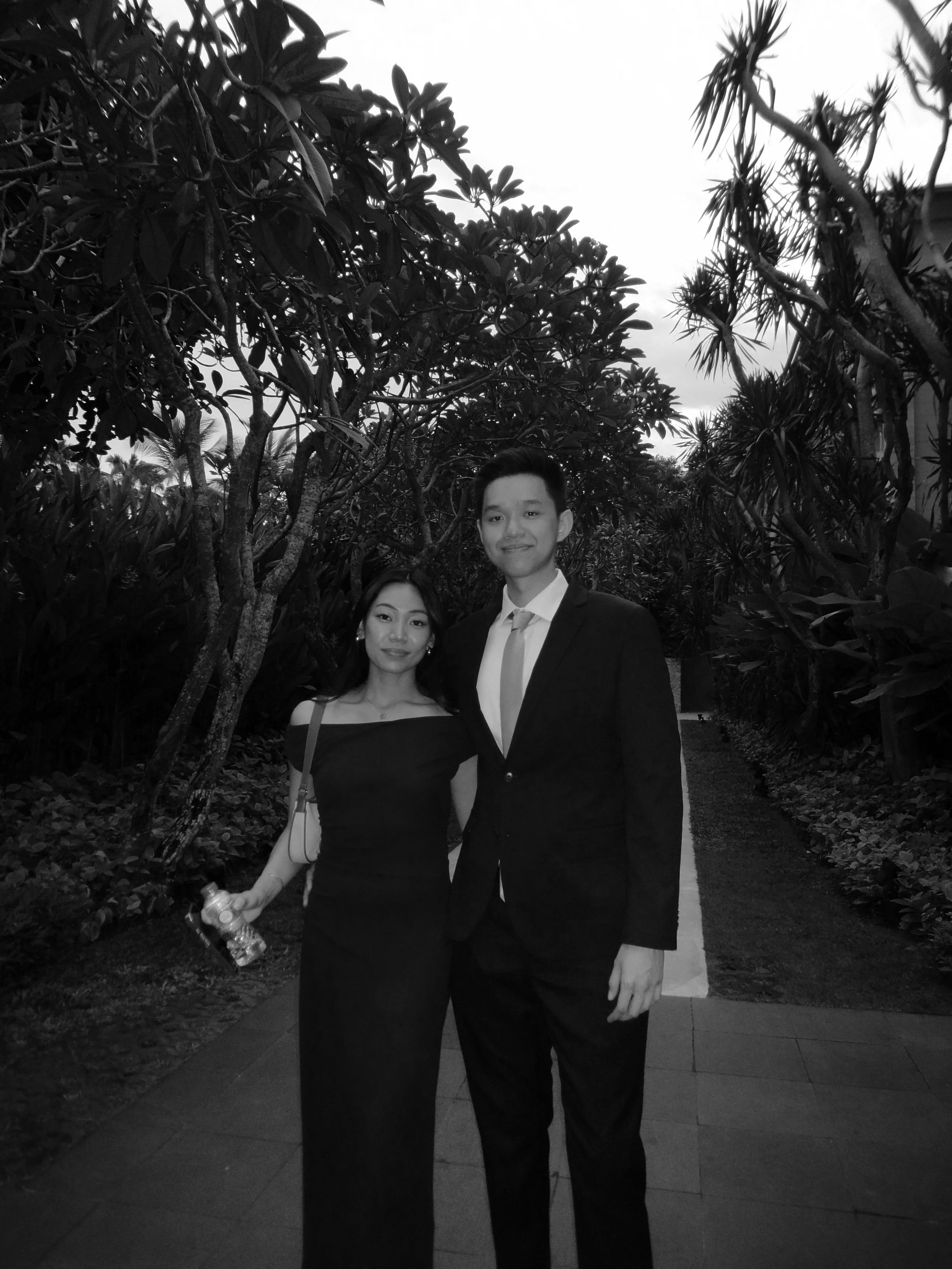 A black and white photo of a young woman and man in formal attire standing outdoors on a pathway surrounded by trees and plants. The woman is holding a water bottle and has a shoulder bag, while the man is wearing a suit and tie.