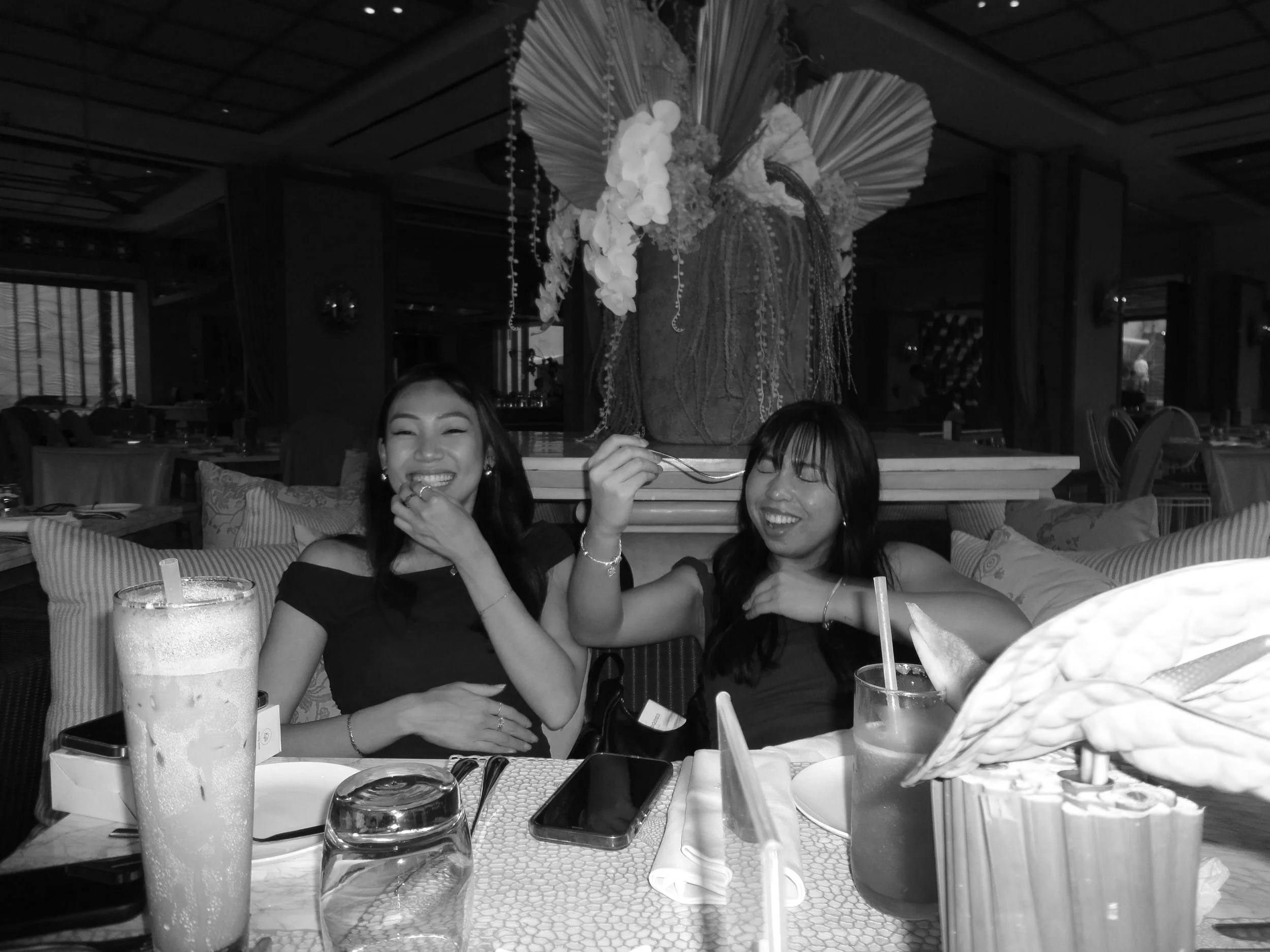 Two women sitting at a restaurant table laughing, with drinks and phones in front of them, and a large floral arrangement behind.