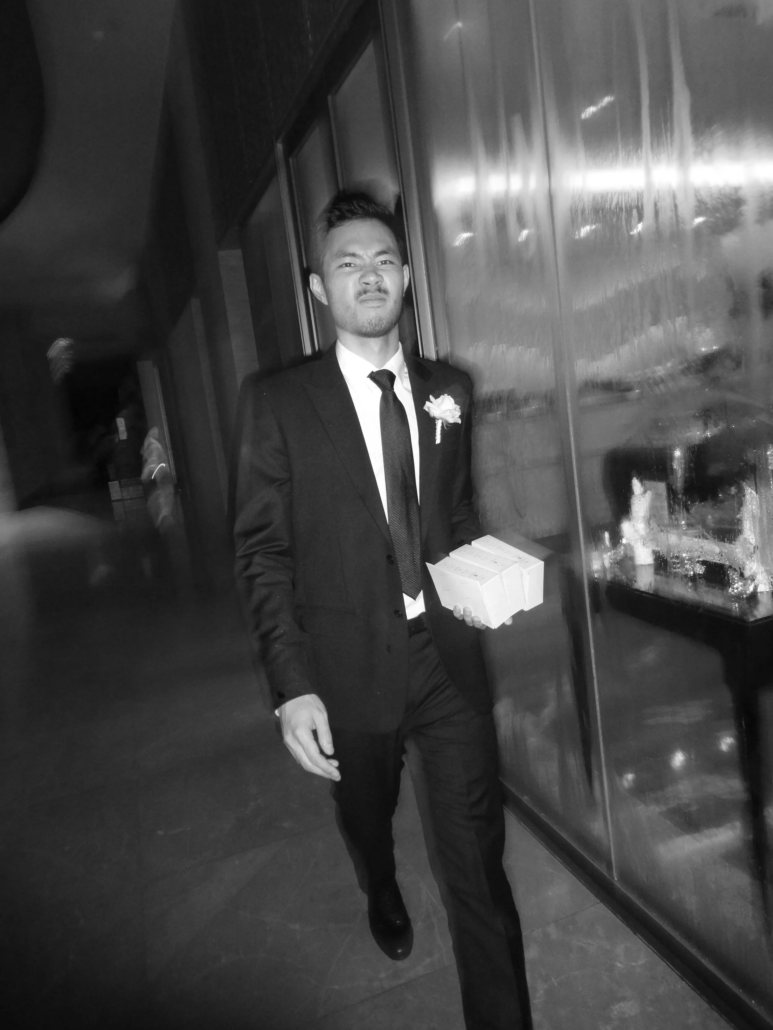 A young man in a suit and tie with a flower boutonniere, holding small boxes, walking indoors past a glass display case, with a skeptical facial expression.