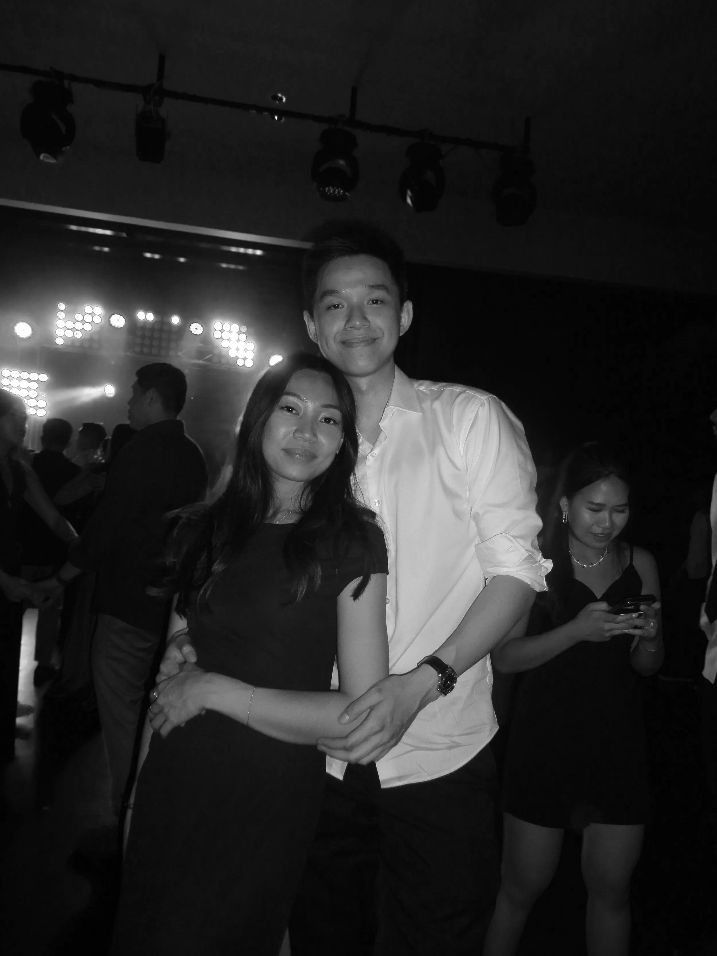 A black and white photo of a young couple smiling and embracing at a party or event, with other guests in the background.